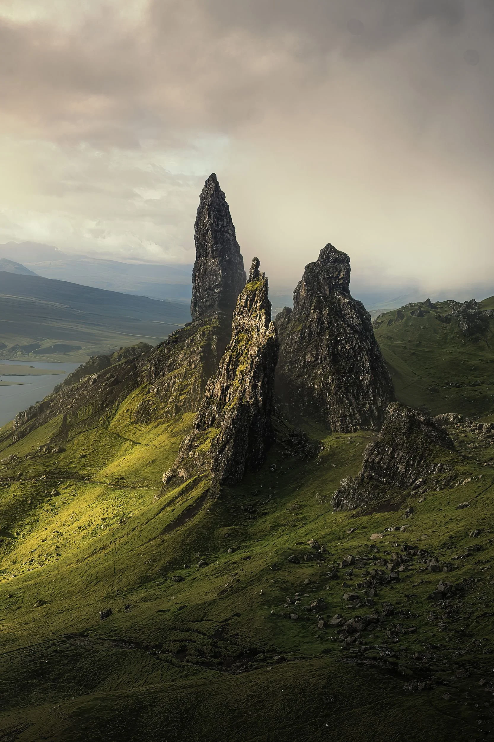 Three tall, dark rocky formations rising from a green hillside under a cloudy sky.