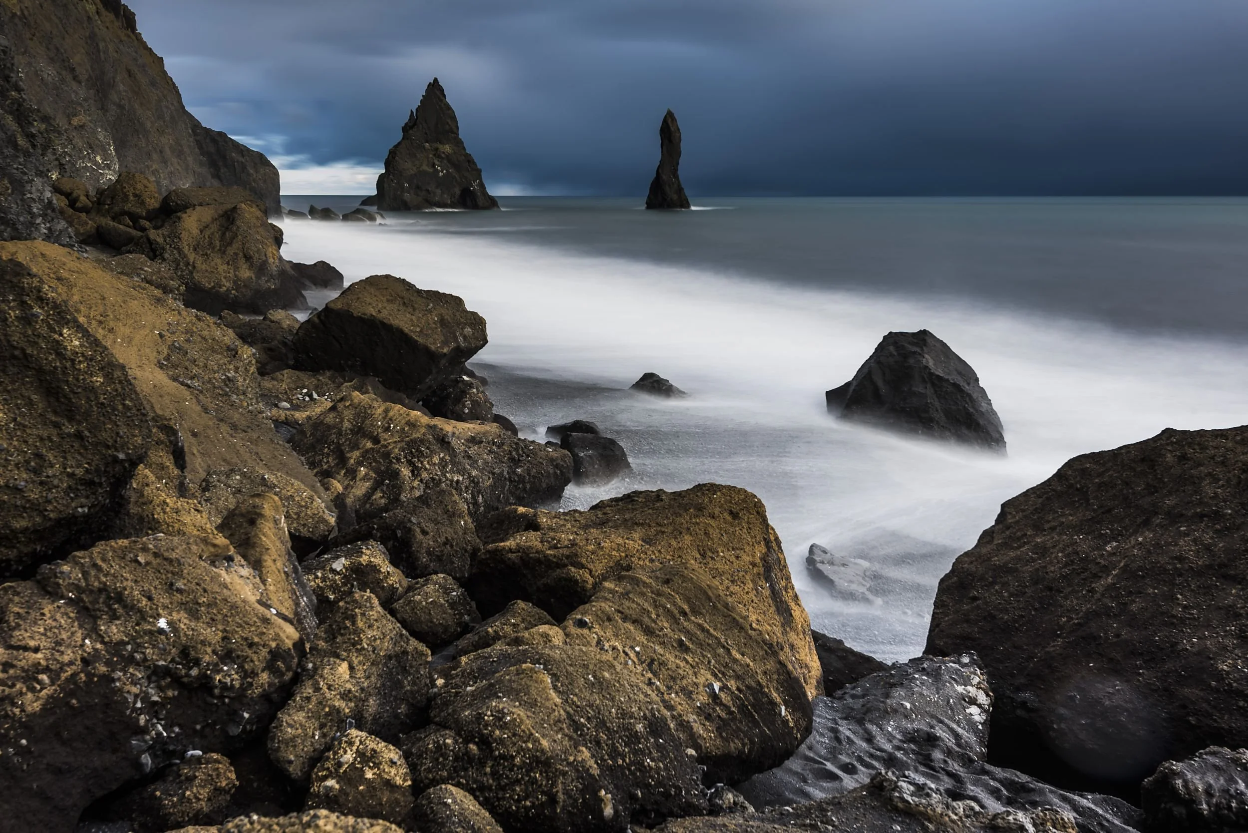 Rocky coastline with large boulders in the foreground and jagged sea stacks in the distance under a dark cloudy sky.