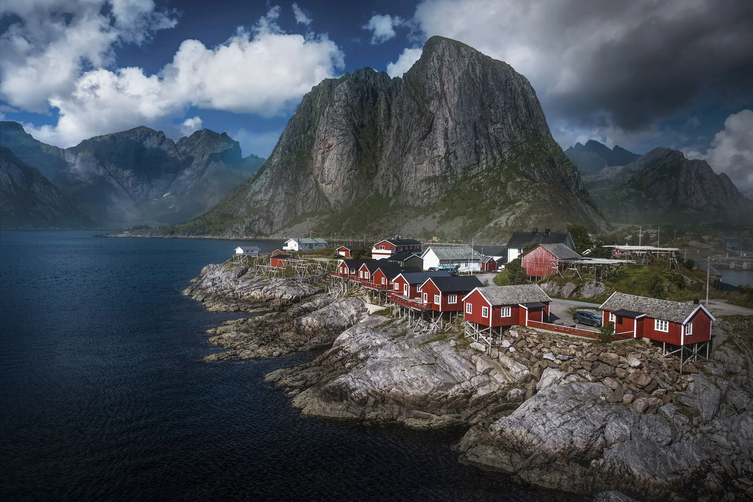 Red wooden houses on stilts along rocky shore with mountain cliffs and fjord in the background under cloudy sky.