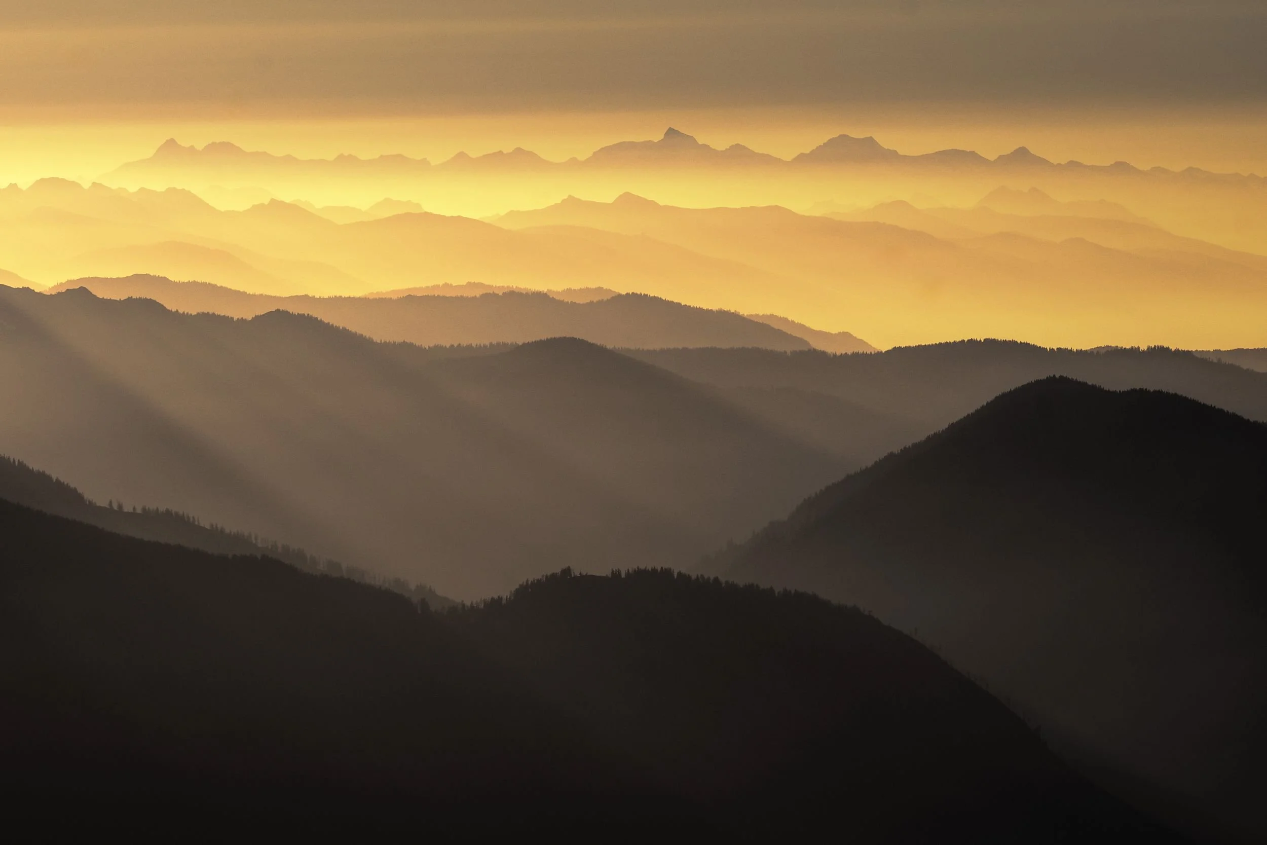 Multiple layers of mountain ranges at sunrise or sunset, with sunlight casting a golden glow and rays penetrating through the clouds.