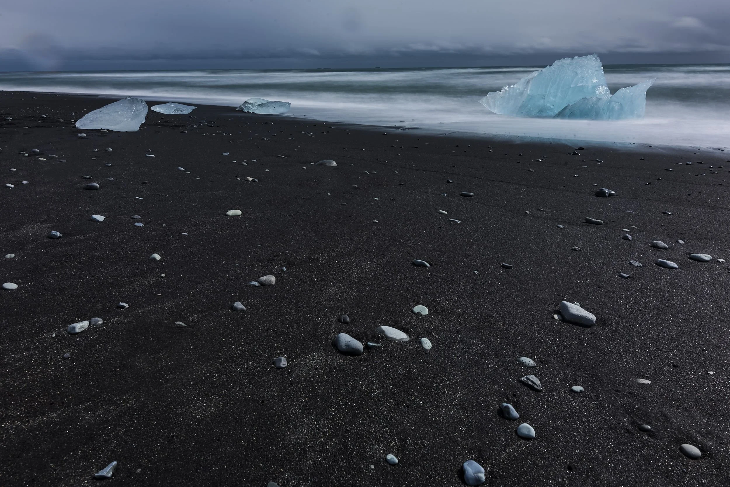 Dark sandy beach with scattered small rocks and pebbles, with large chunks of ice and icebergs washed ashore, under a dark cloudy sky.