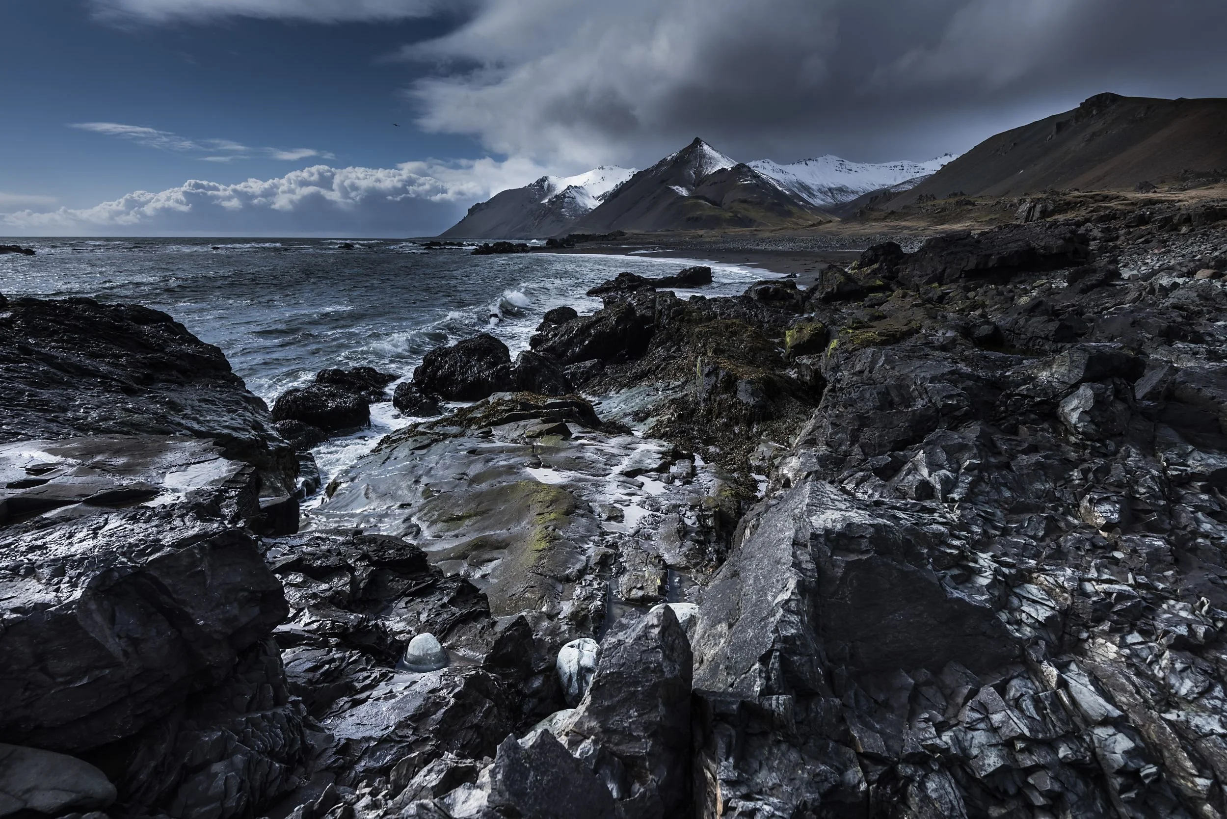 Rocky coastline with turbulent ocean waves and snow-capped mountains under a cloudy sky.