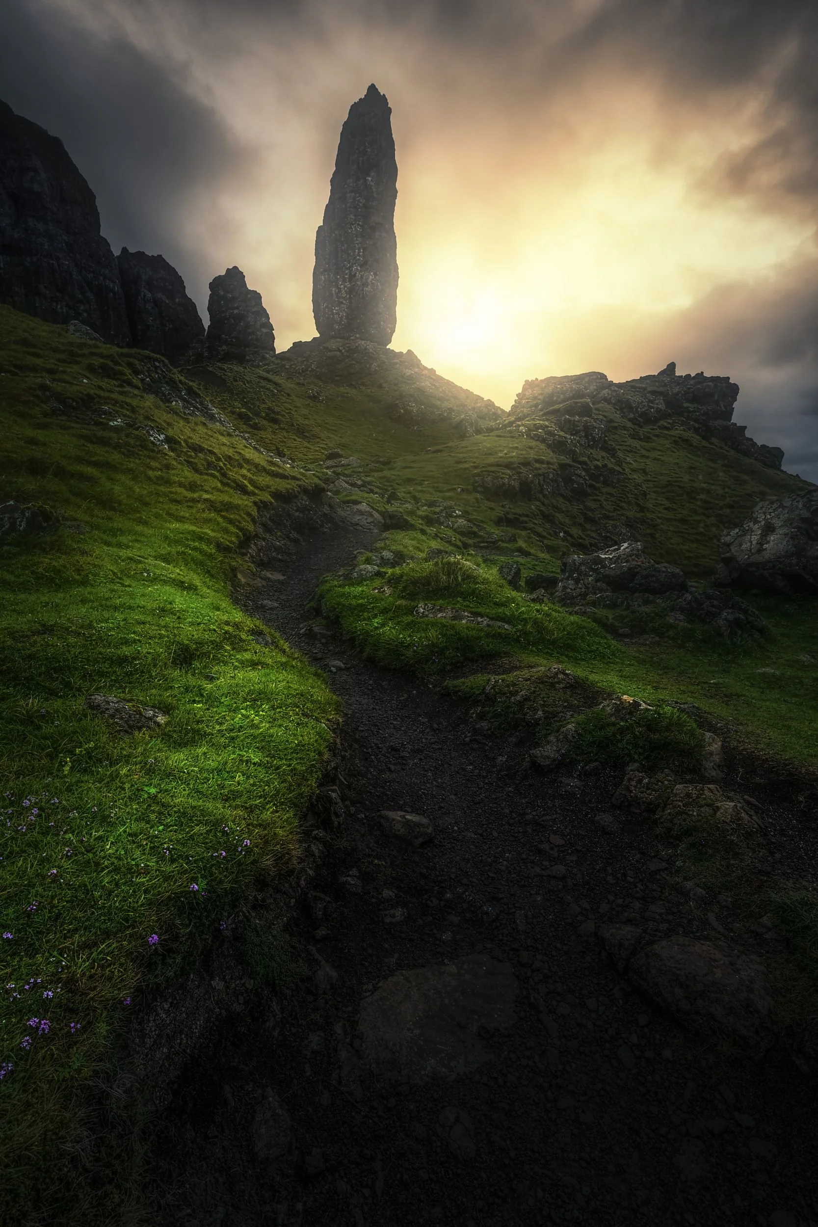 A rugged mountain path leading up to towering rock formations at sunset, with green grassy slopes and a cloudy sky.