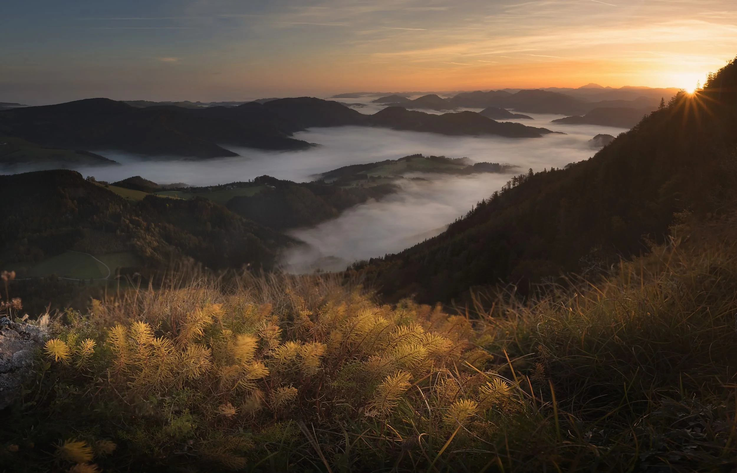 Sunrise over a mountain valley with fog covering the landscape, grassy foreground, and distant mountain ridges.