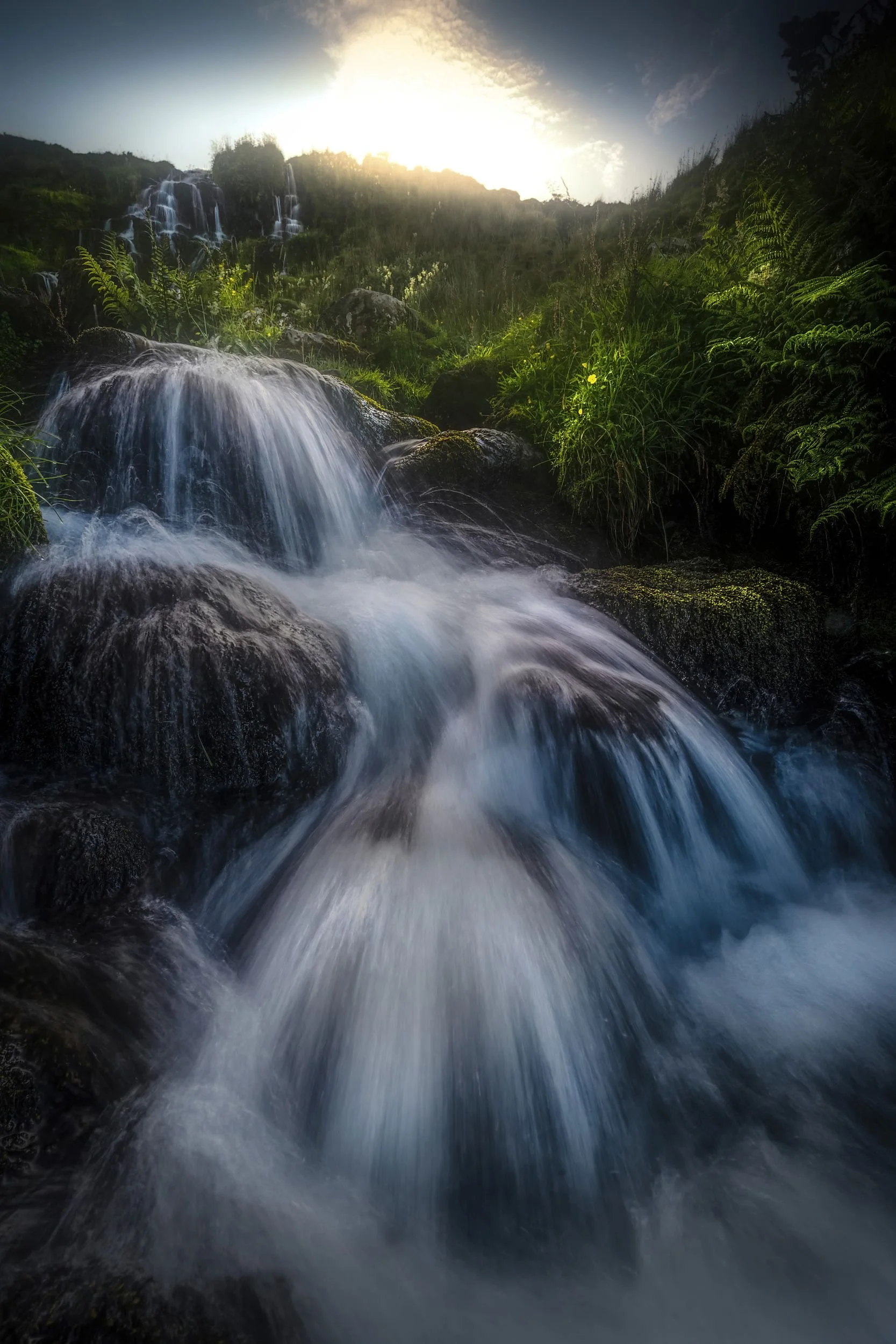 Ein Wasserfall fließt über Felsen in einer grünen Landschaft bei Sonnenuntergang.