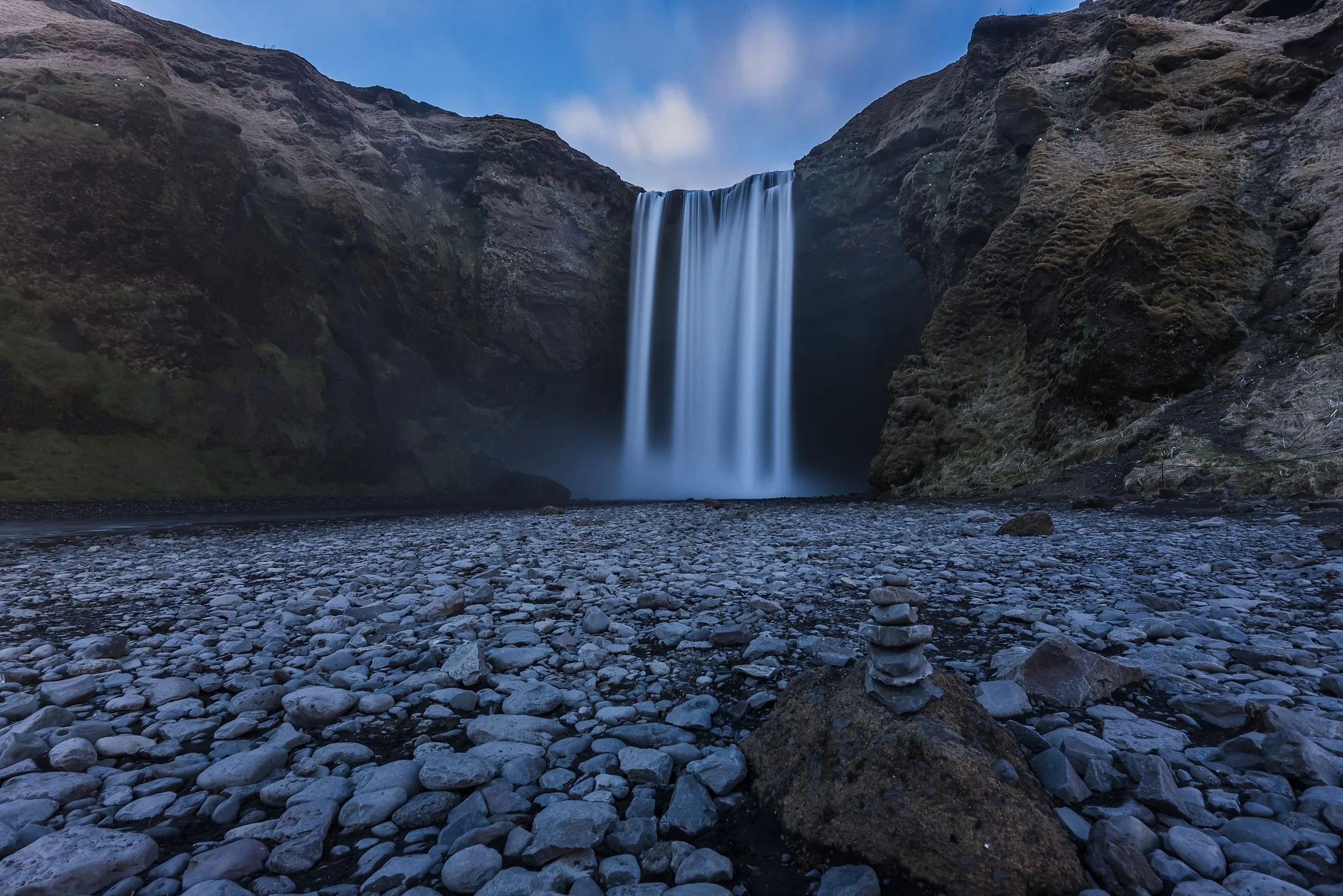 A waterfall cascading into a rocky riverbed, surrounded by moss-covered cliffs under a blue sky.