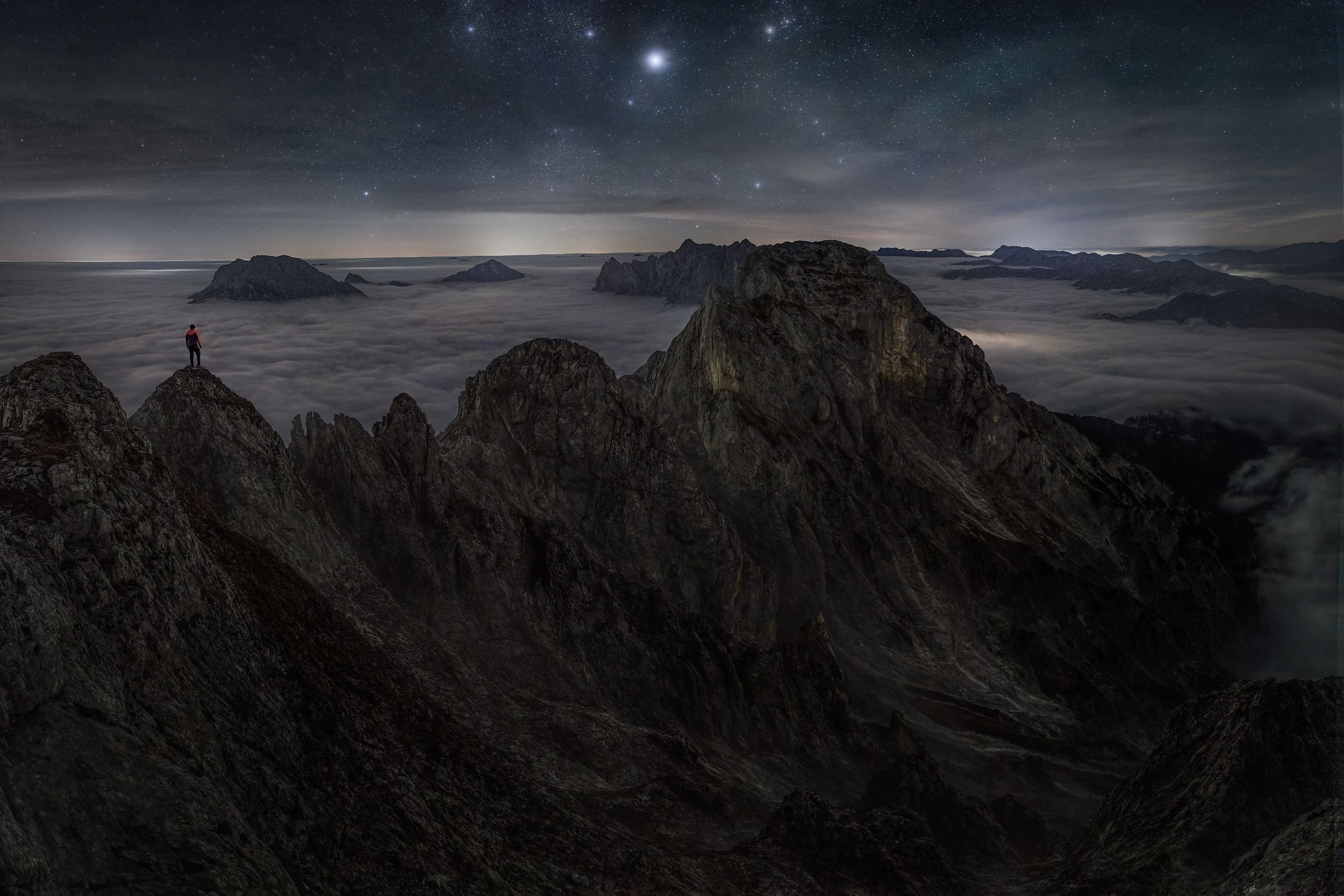 A person standing on a rocky mountain peak at night, overlooking a sea of clouds below and a starry sky above.
