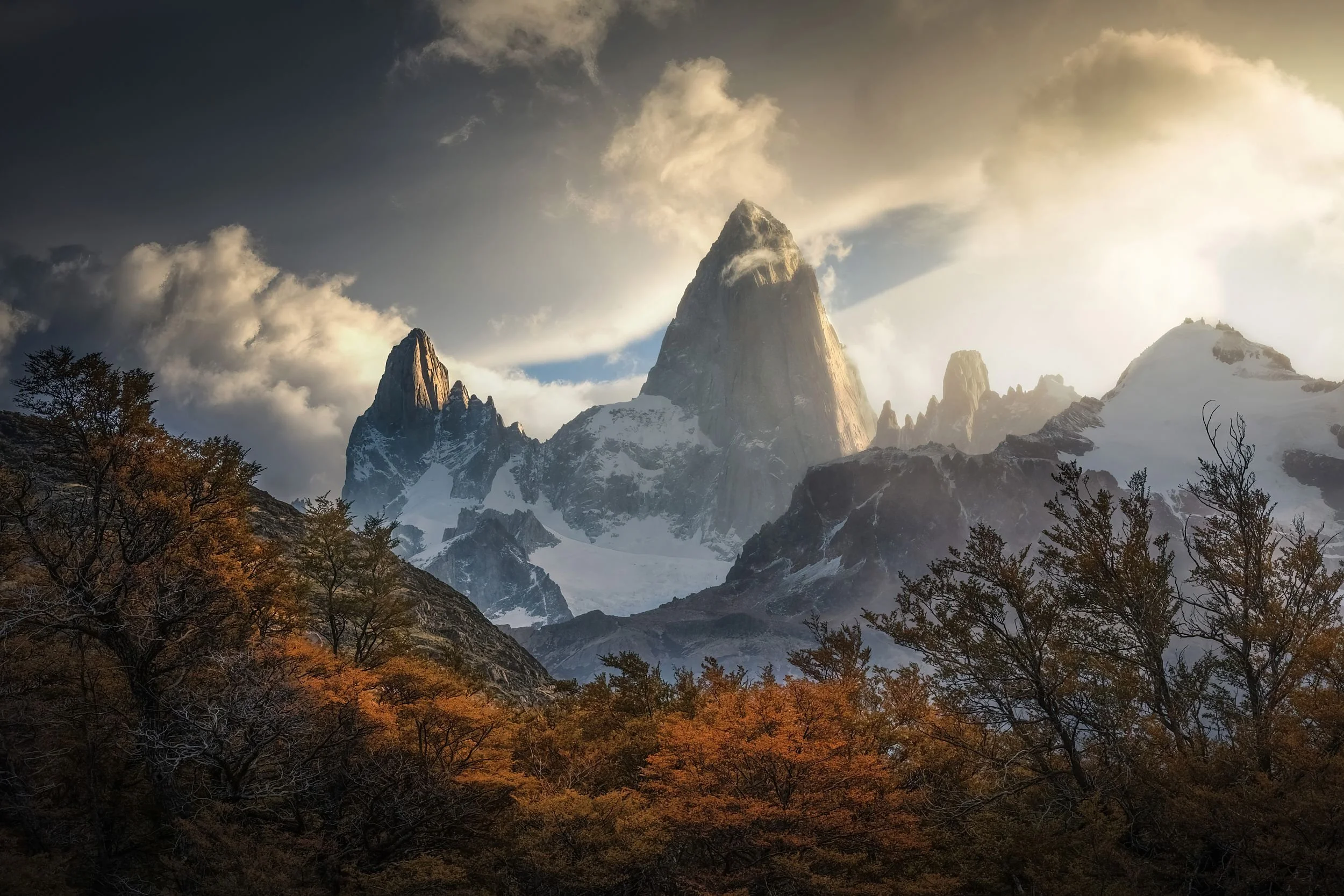 Mountain landscape with tall peaks, some snow-capped, with clouds in the sky and autumn-colored trees at the base.