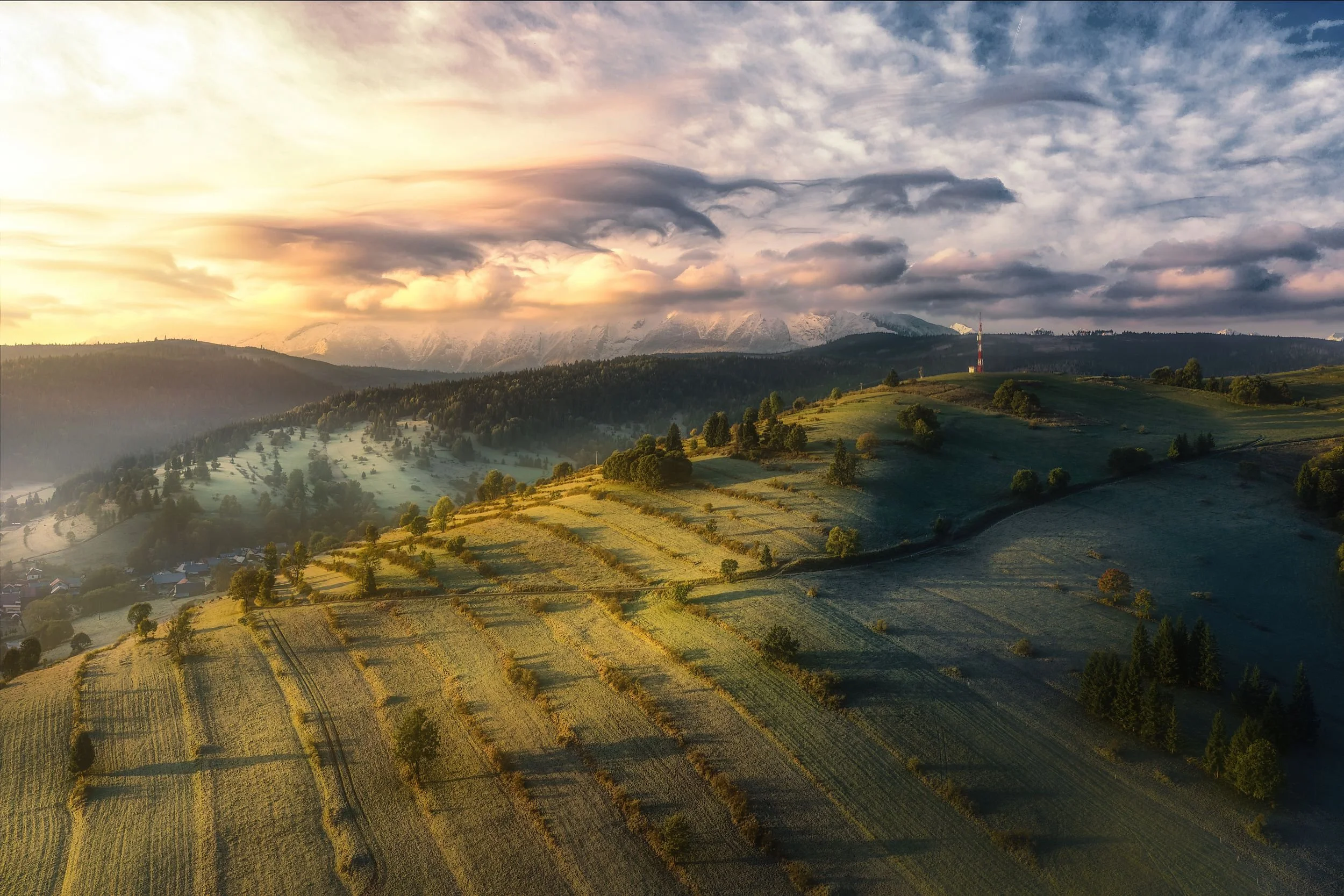 A landscape of rolling green hills with patches of farmland, trees, and a small village, under a sky with clouds and sunlight breaking through, with snowy mountains in the background.