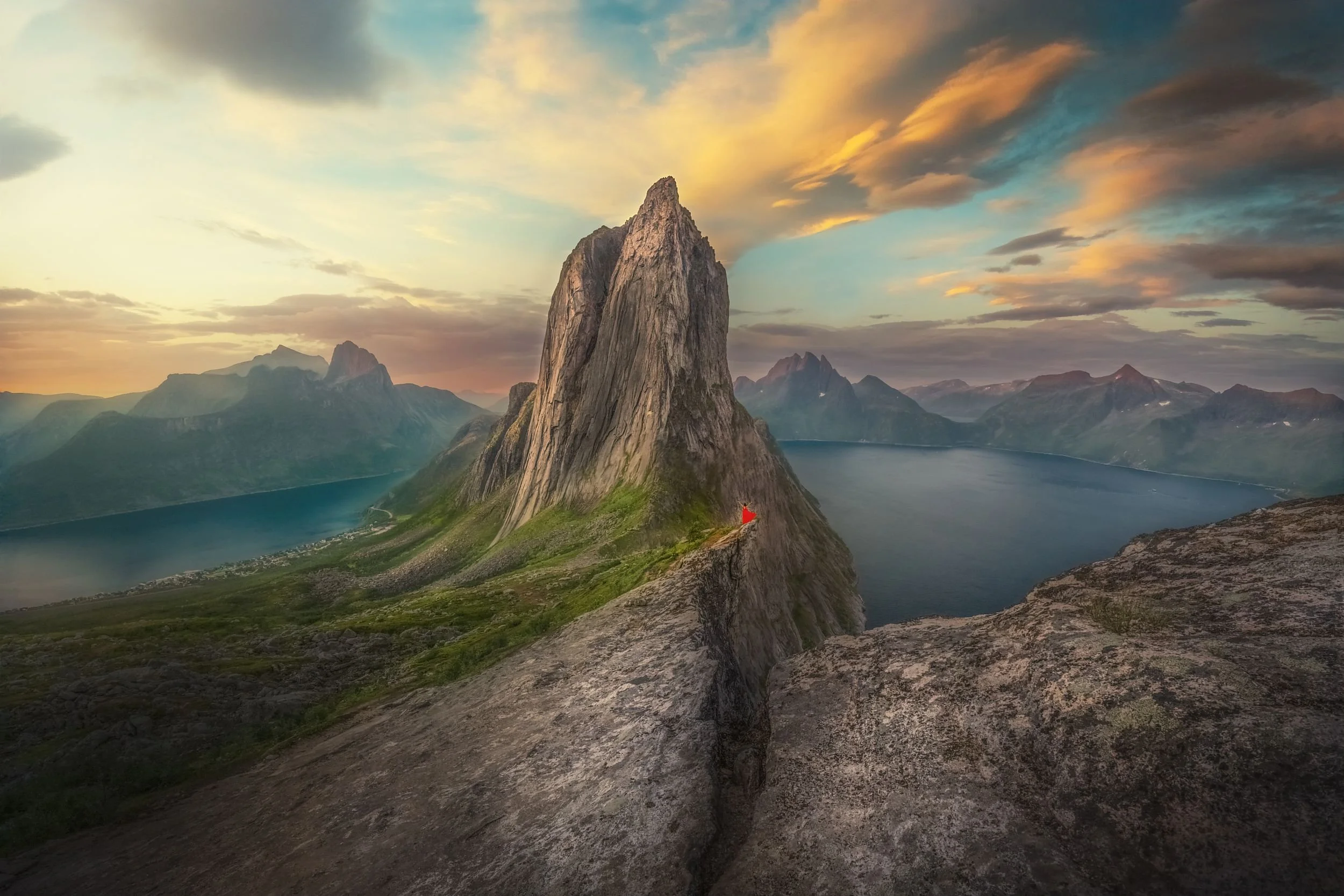 A large rocky mountain with steep slopes overlooking a lake, with a sunset sky and clouds in the background.