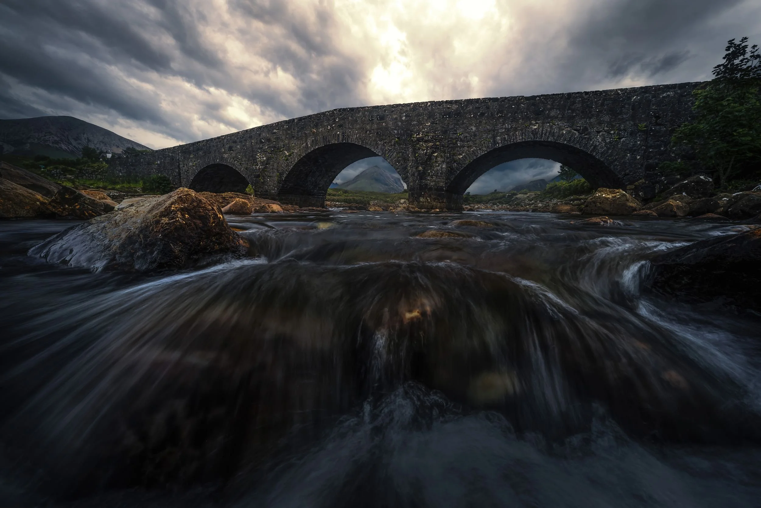 Ein steinerne Brücke mit mehreren Bögen überquert einen Fluss, im Hintergrund Berge und ein bewölkter Himmel.