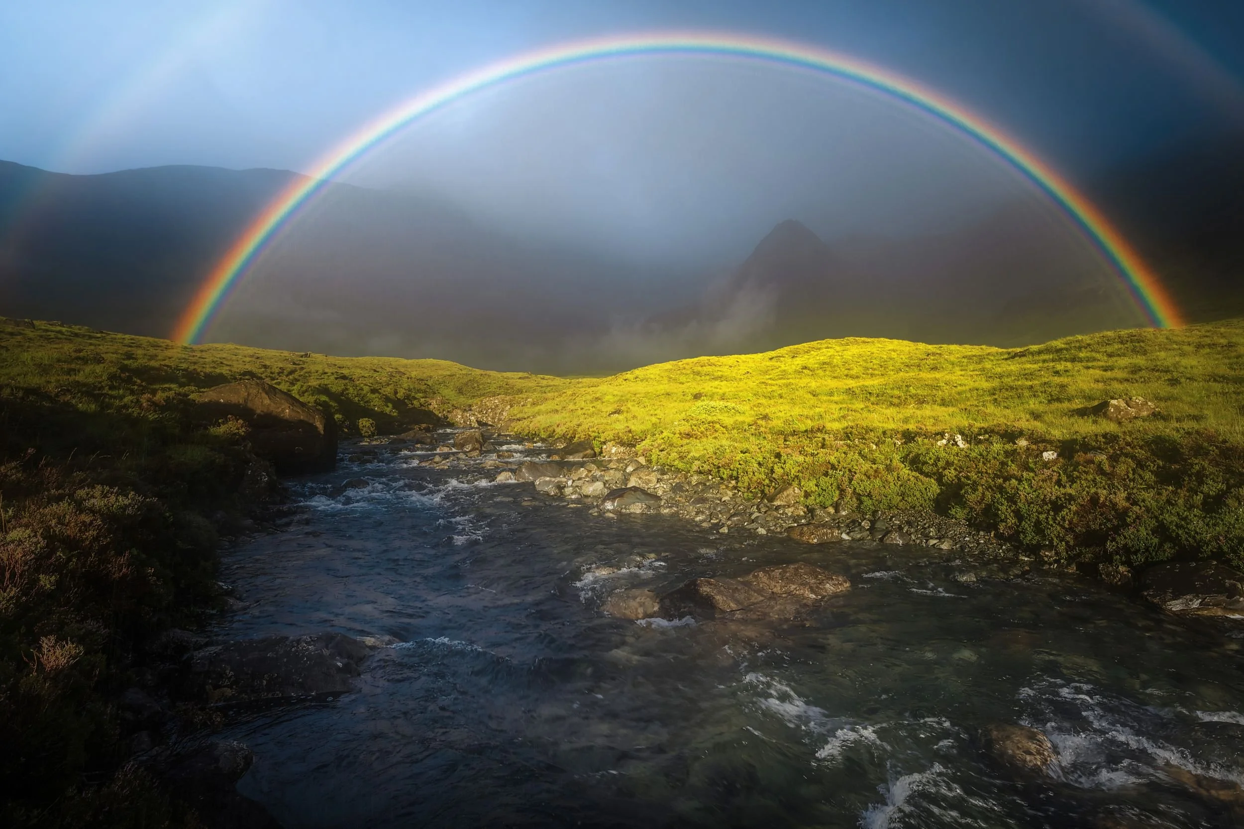 Mehrfacher Regenbogen über einer grünen Landschaft mit Hügeln und einem Fluss im Vordergrund, bei bewölktem Himmel.