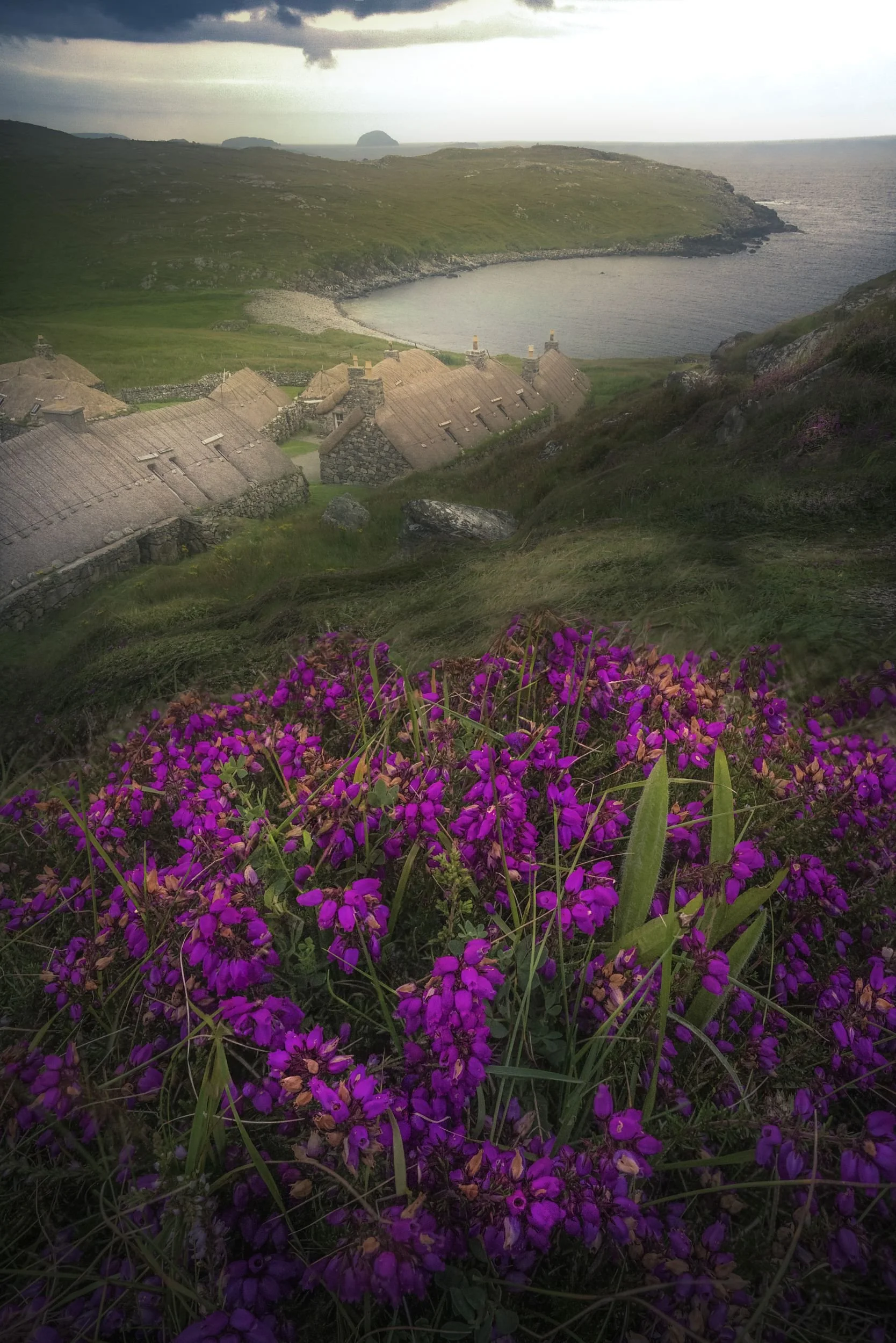 Blick auf eine grüne Hügellandschaft mit einem kleinen See im Vordergrund, eine Reihe von alten Steinhäusern mit Schilfdächern, im Hintergrund Meer und Felsen, im Himmel dunkle Wolken.