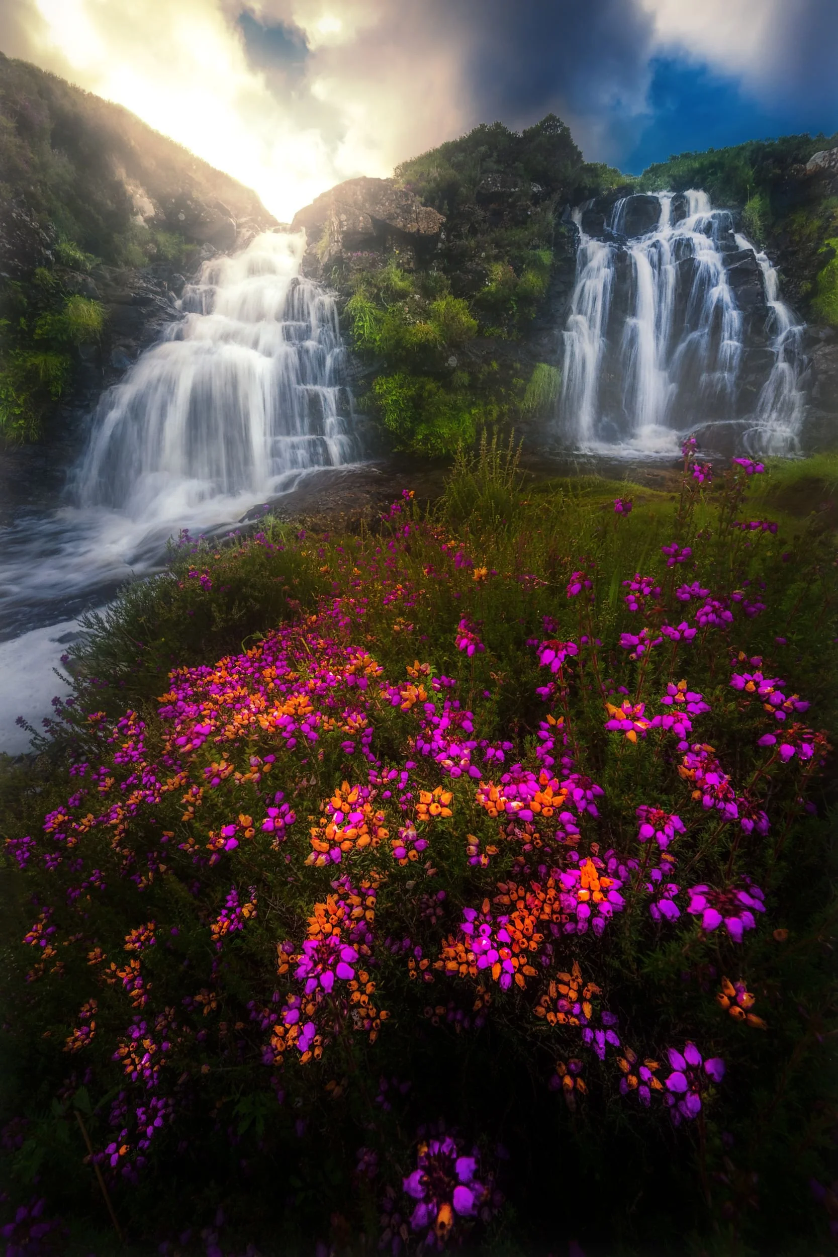 Mehrere Wasserfälle in einer grünen hügeligen Landschaft bei bewölktem Himmel, bunte Blumen im Vordergrund.