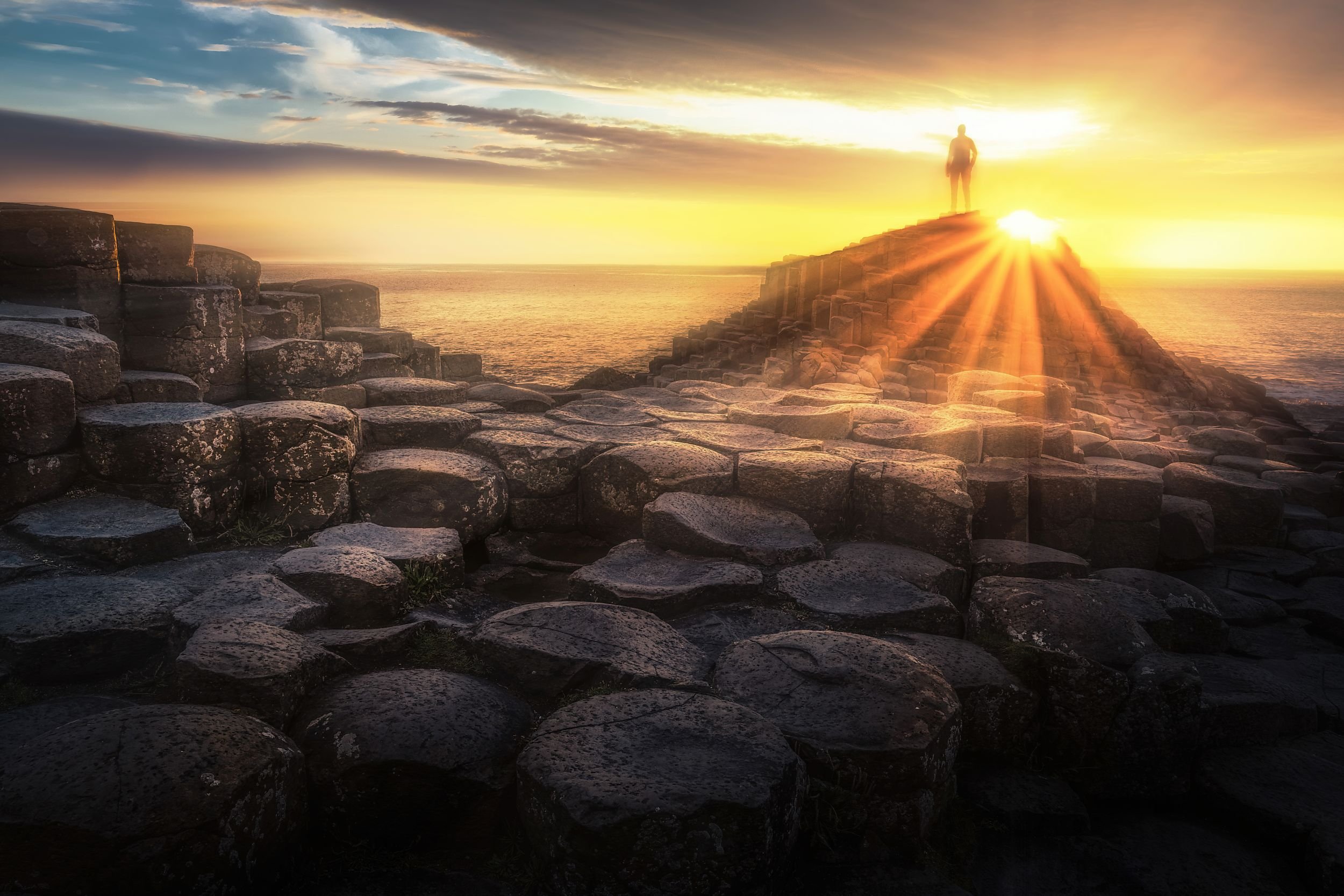A person standing on a stepped volcanic rock formation near the ocean during sunset, with the sun shining brightly behind them and illuminating the sky with warm colors.