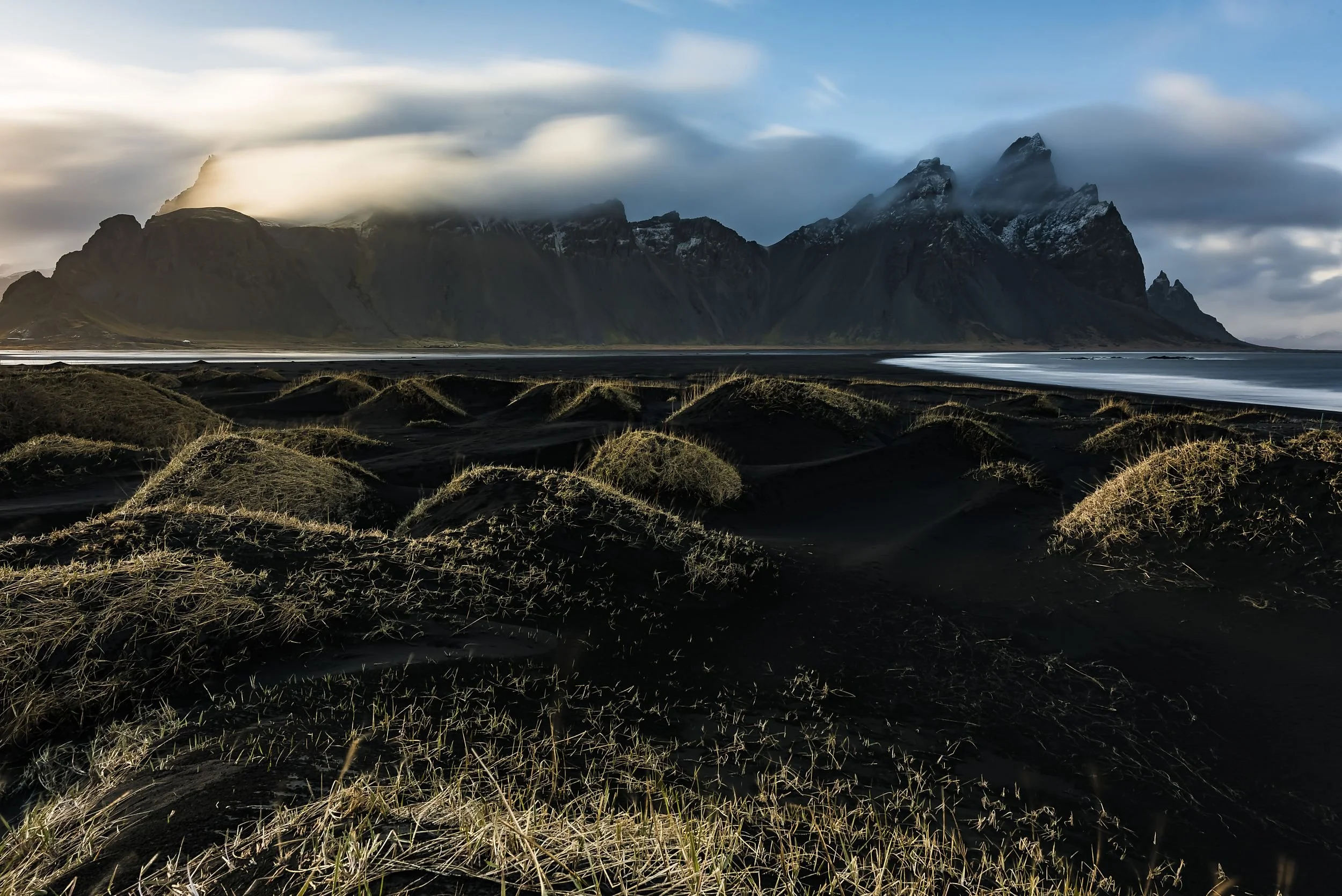 Dark volcanic beach with black sand and patches of grass, rugged mountains in the background partially covered by clouds, and an overcast sky.