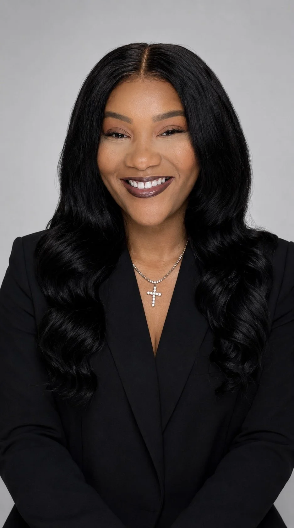 A woman with long black wavy hair, wearing a black blazer, a silver cross necklace, and smiling at the camera against a gray background.