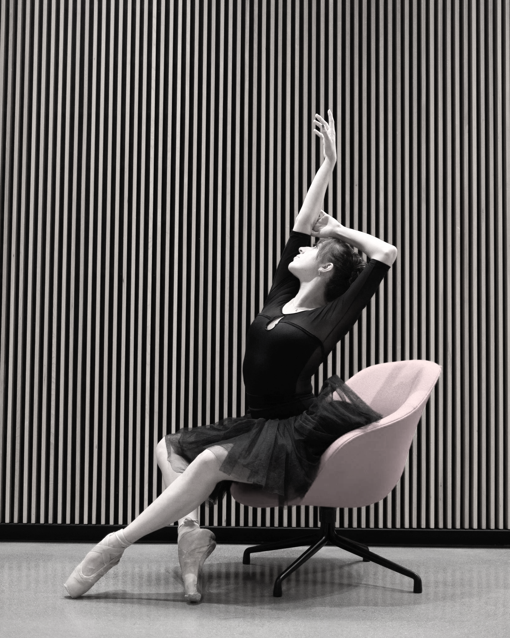 A dancer in a black tutu and pointe shoes luxuriates in a chair, arms yawing upwards.