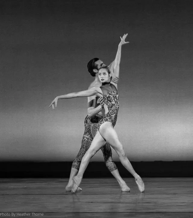 A dancer on pointe in a huge fourth position, supported by another dancer standing behind her, and looking away from her.