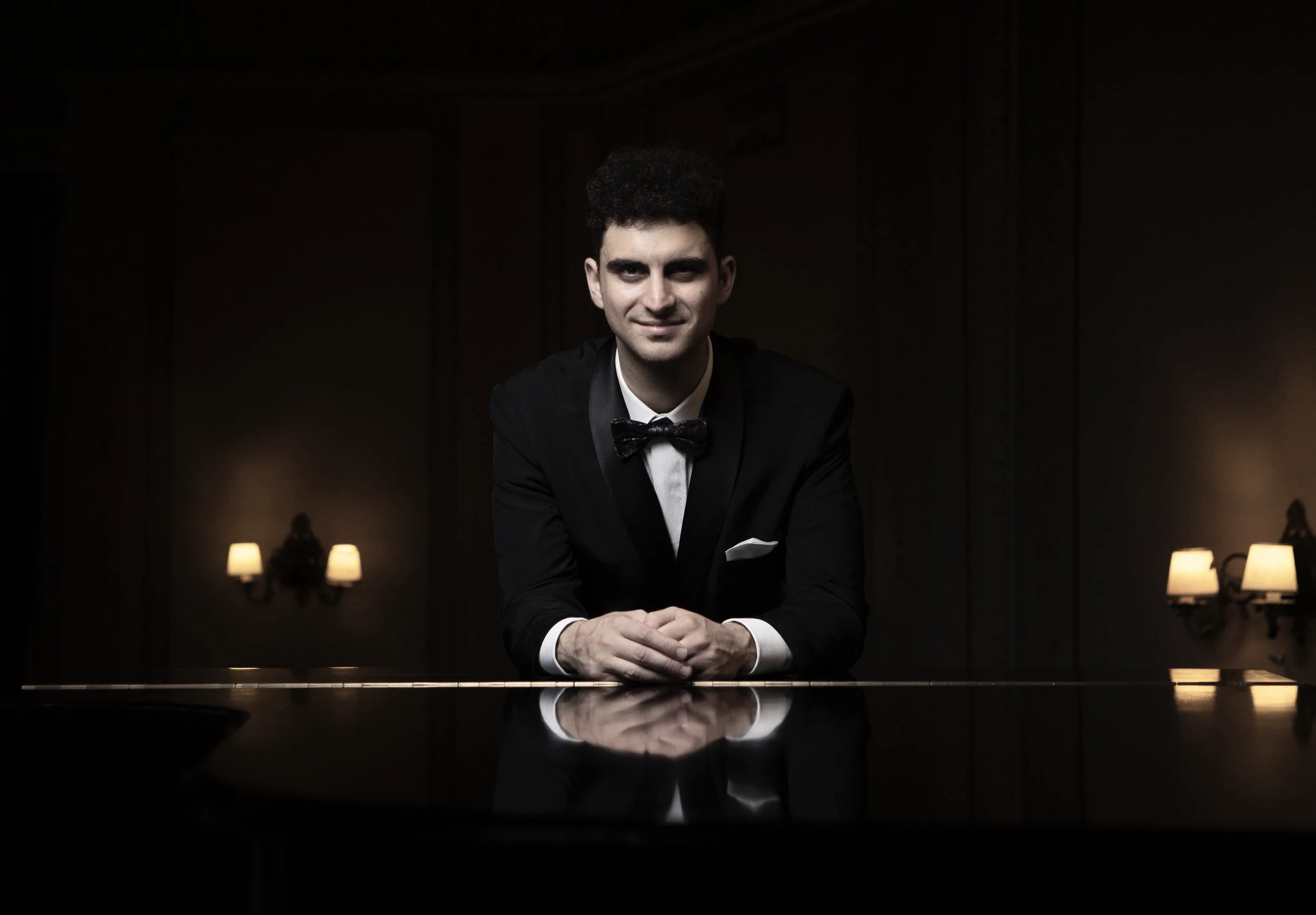 A man in a tuxedo sits facing the camera with his hands folded on top of a piano.