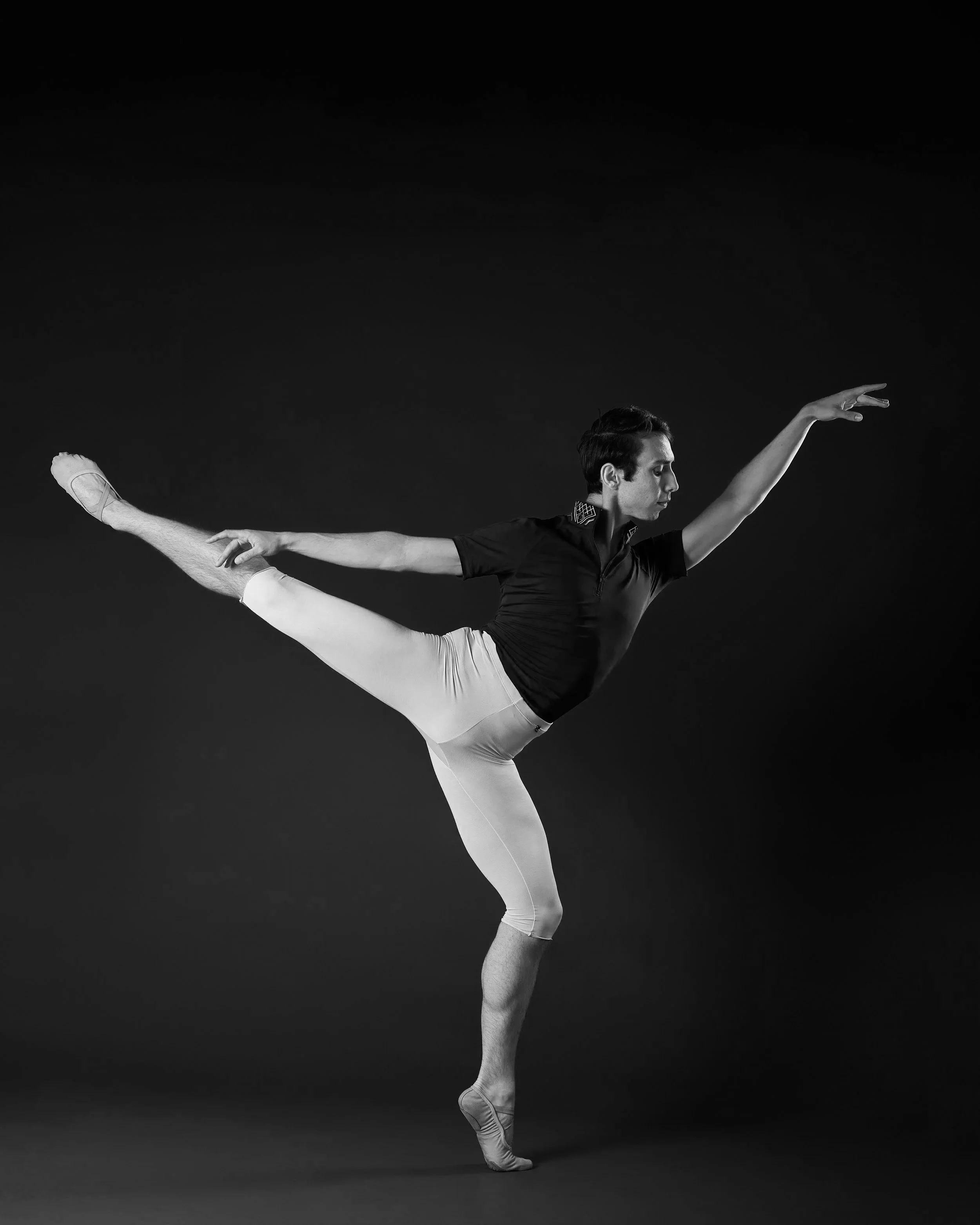 A dancer in a dark top and white pants holds a first arabesque on a bent knee demi pointe.