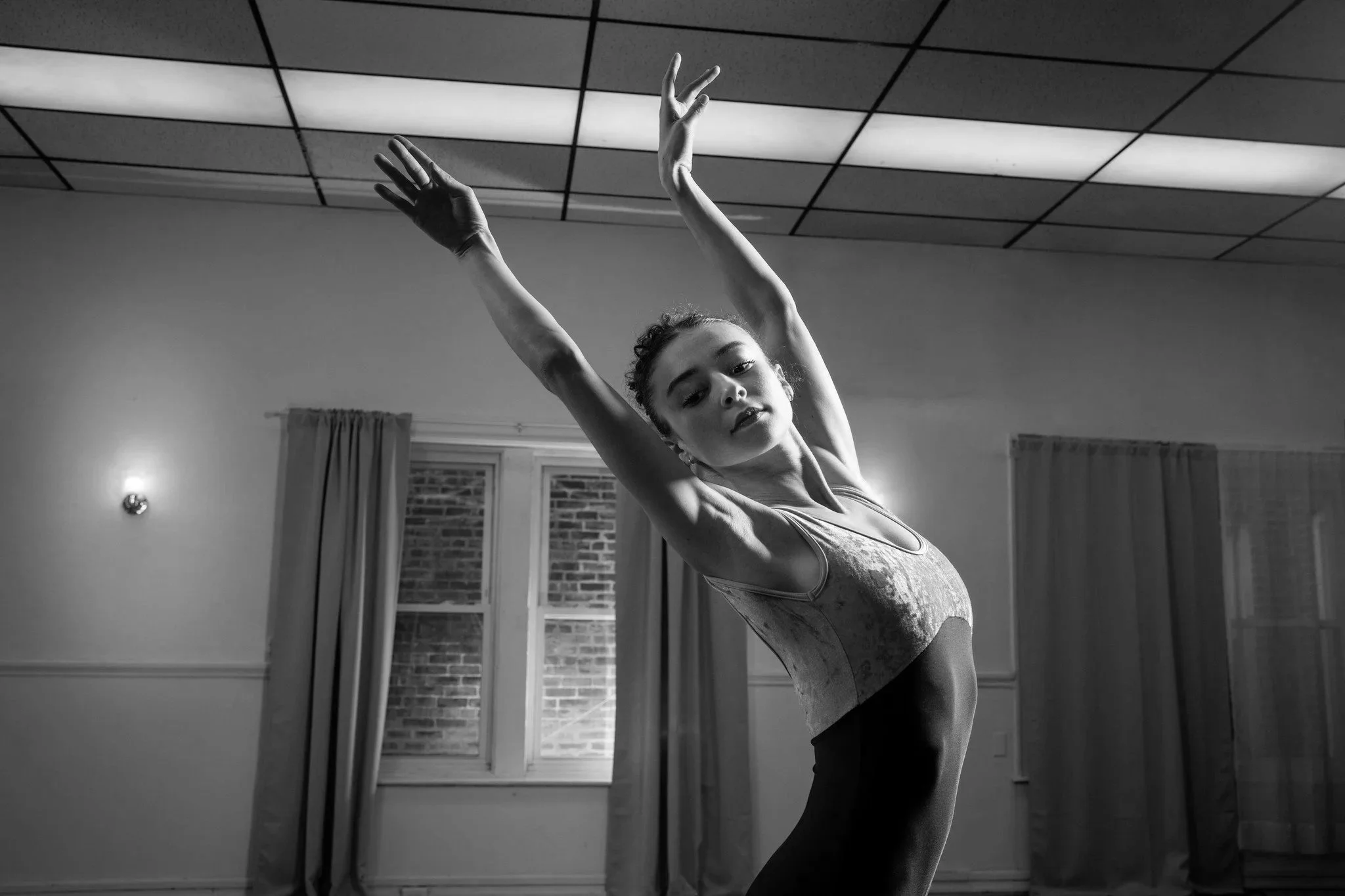 A dancer in a grey blue leotard arches her back, eyes downcast and palms facing out.