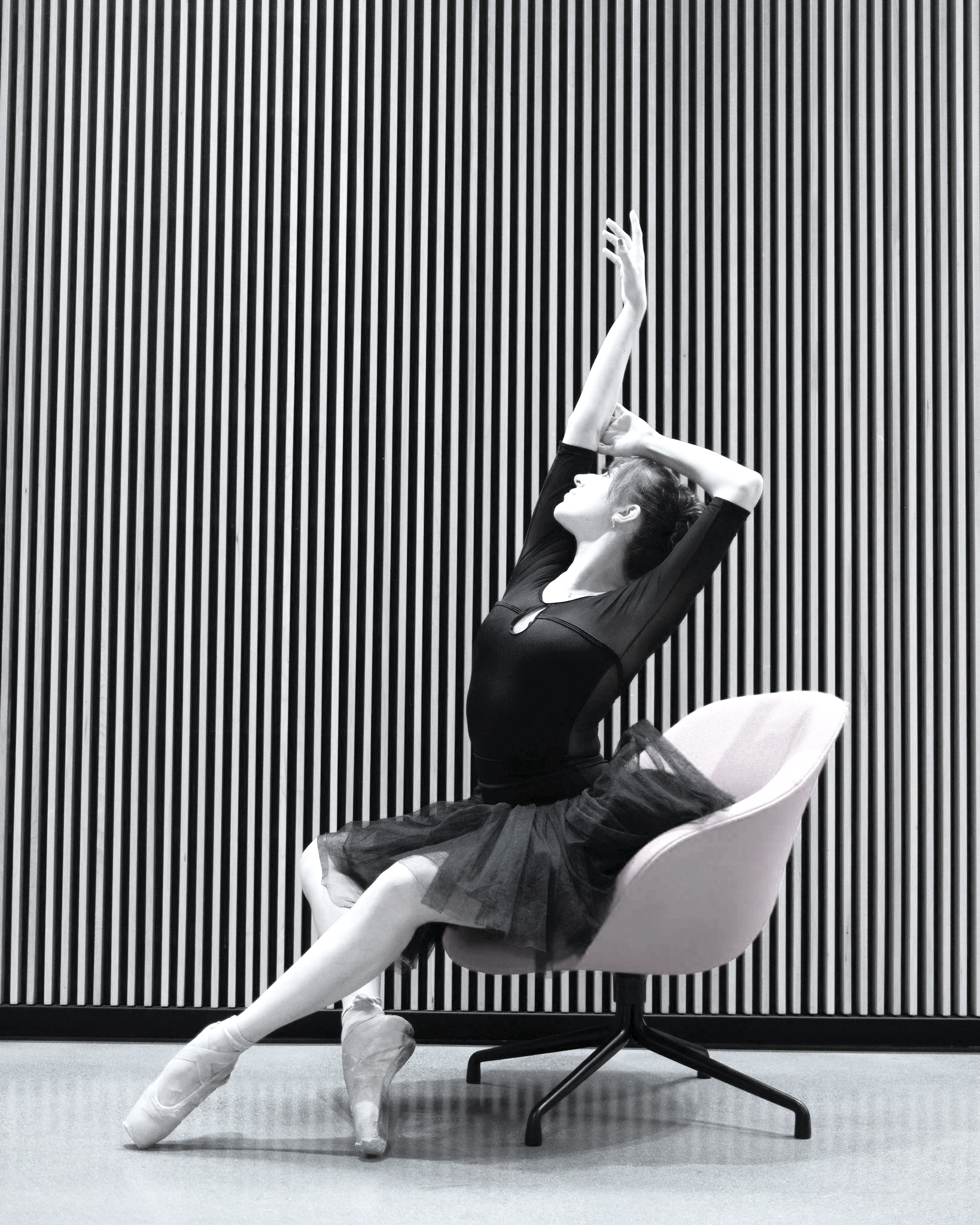 A dancer in a black tutu and pointe shoes luxuriates in a chair, arms yawing upwards.