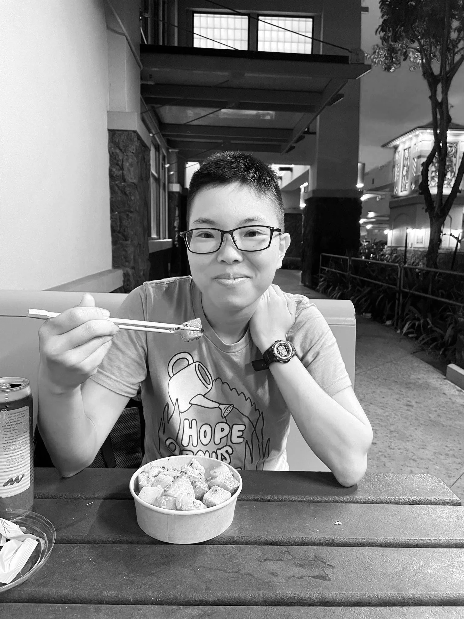A dancer smiles at the camera, holding chopsticks and sitting at an outdoor cafe.