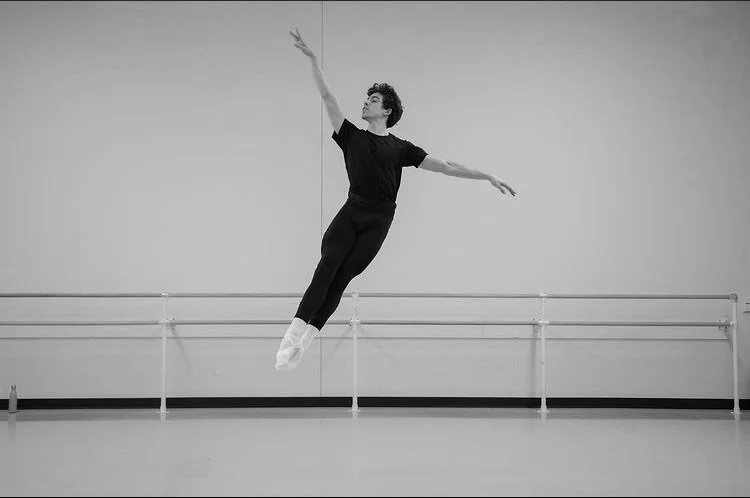 A young male dancer soars through the air, legs together and arms widespread.