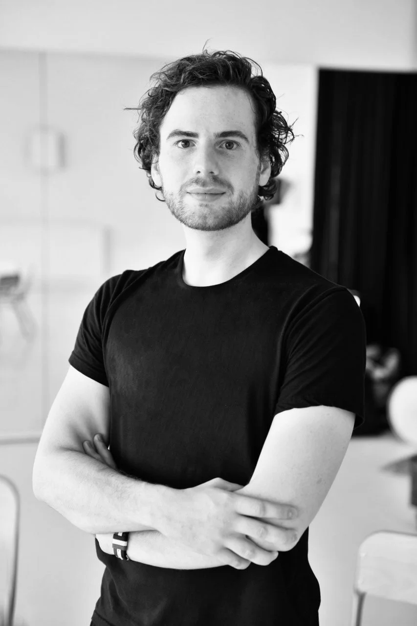 A young man smiles confidently at the camera, arms crossed and Belgian flag bracelet showing on his wrist.