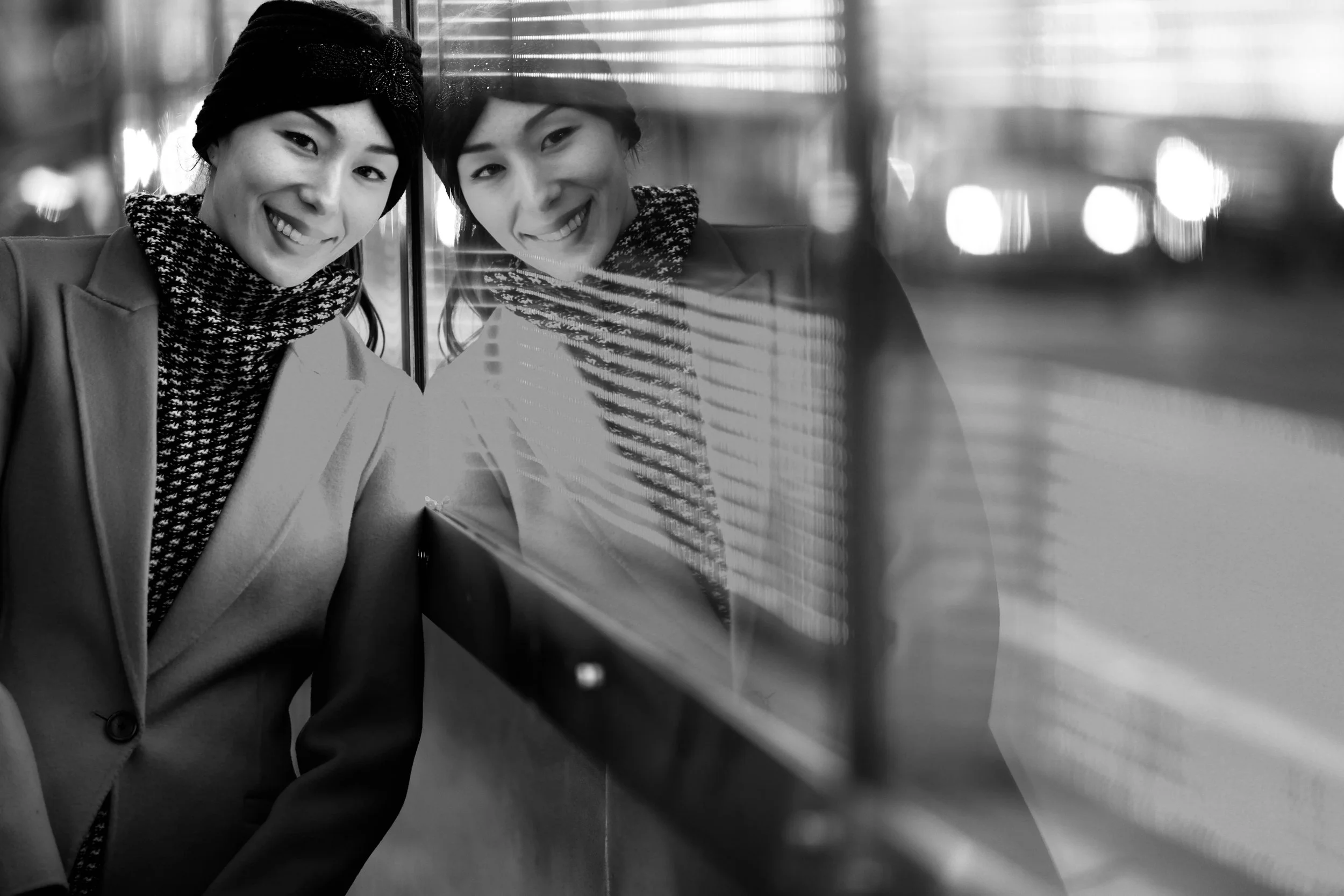A sweetly smiling woman in a headband and smart winter coat leans on a reflective window so it seems there are two of her.