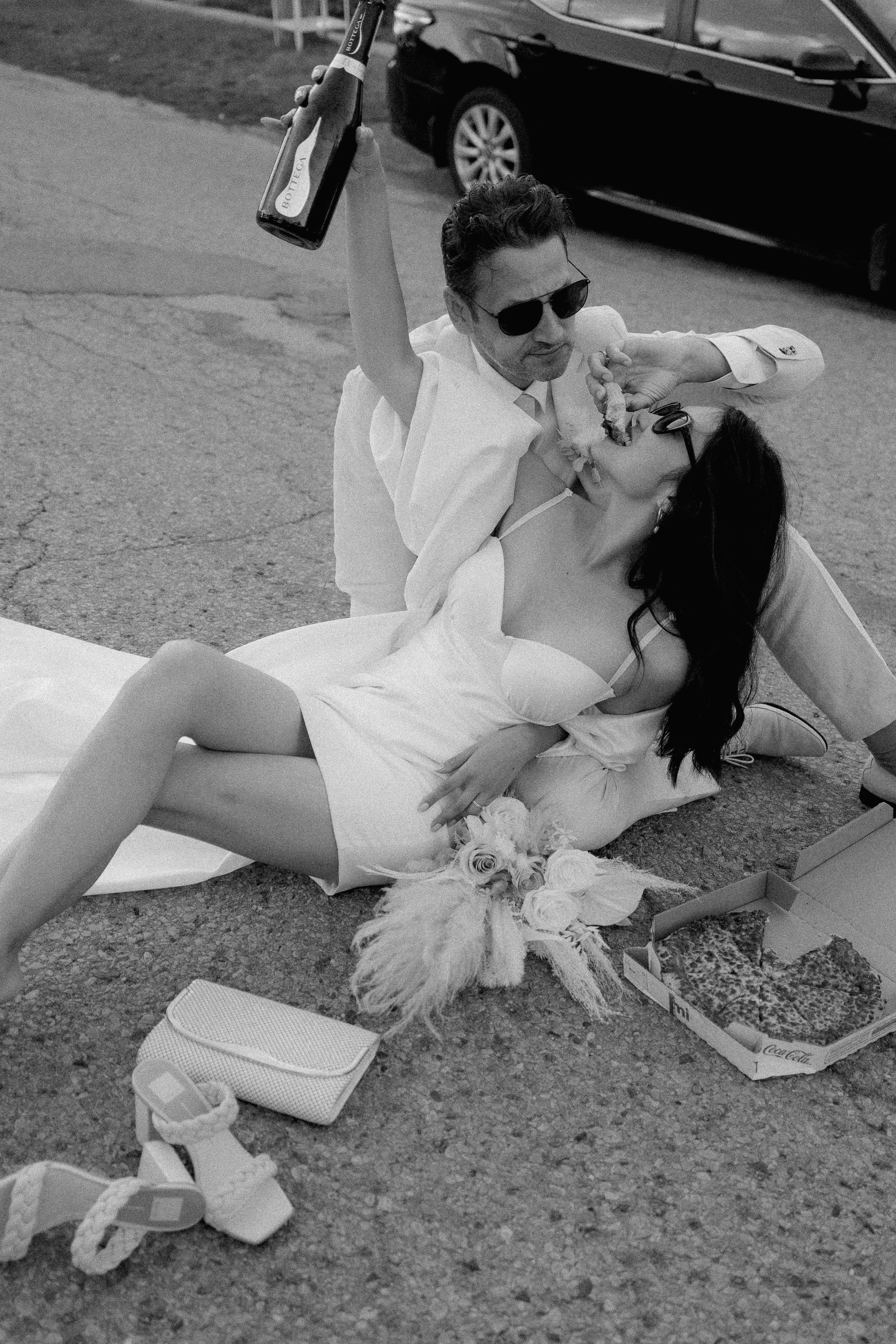 groom feeding bride pizza while bride lays on his lap and holds champagne bottle in hand