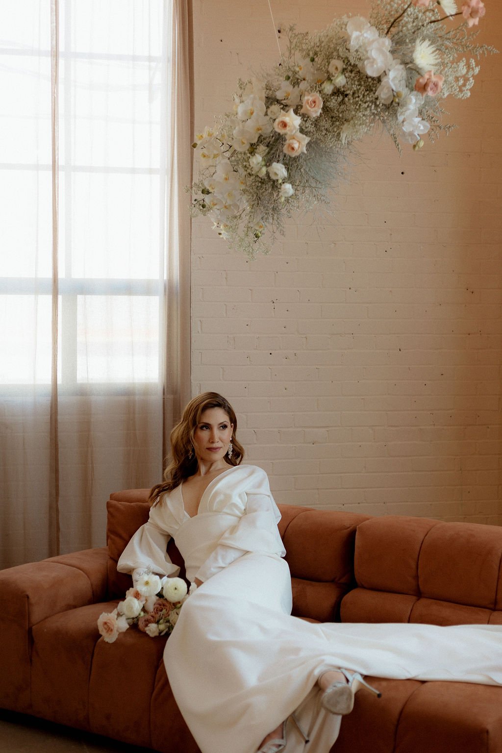 bride sitting on red orange couch while flower arch above her while holding onto her flower bouquet