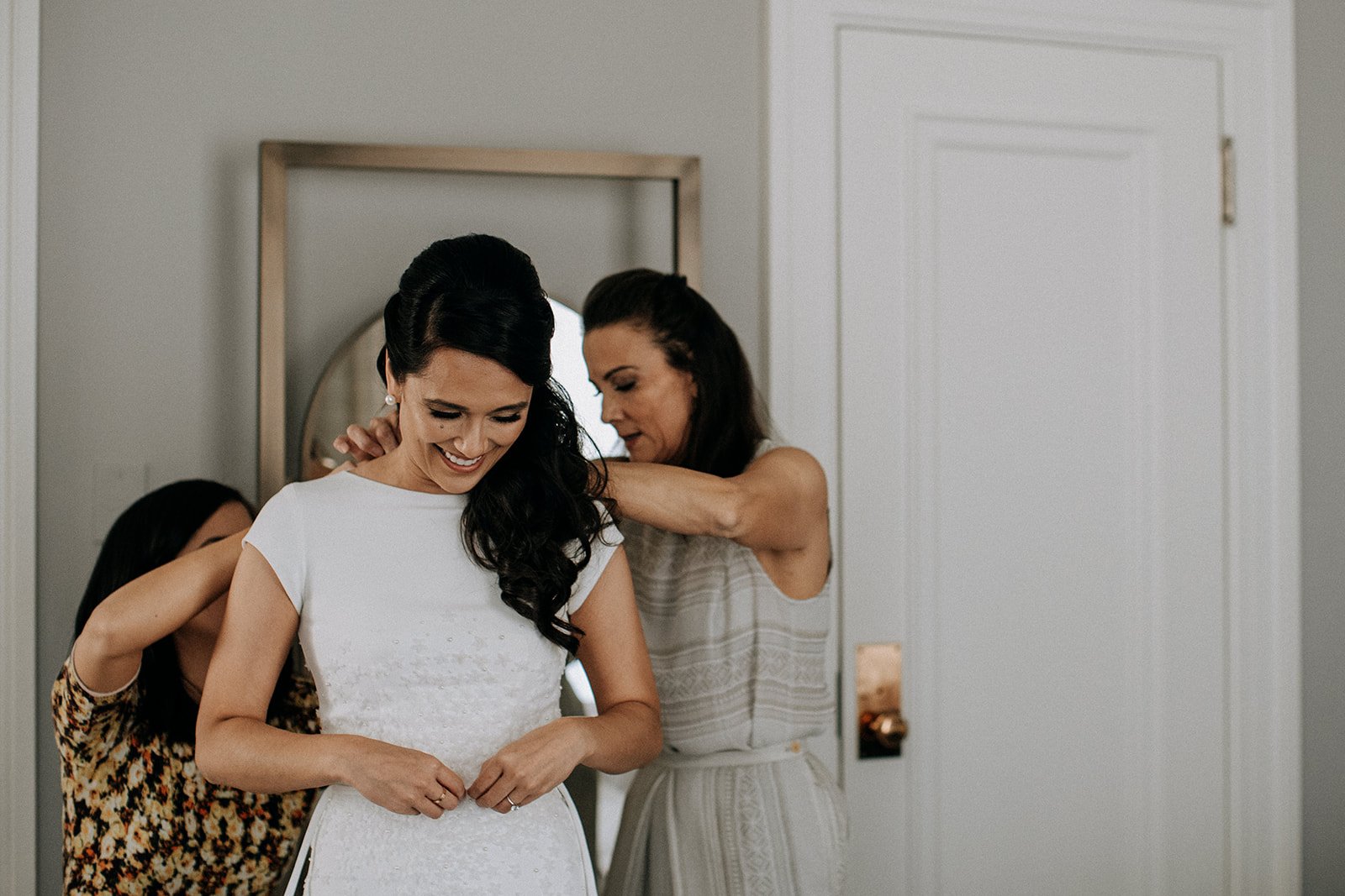  two women helping a bride button her dress 
