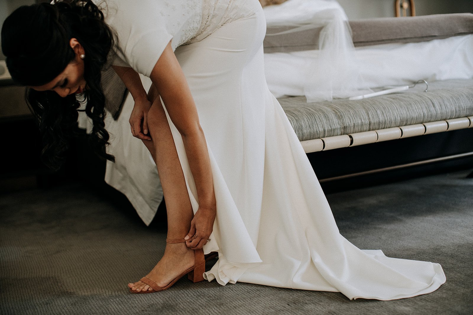  bride putting on her shoe in front of bed 