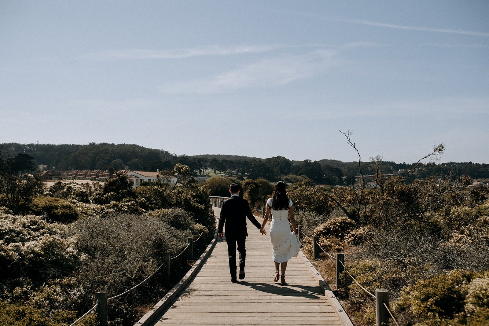  bride and groom walking on a board walk with foliage surrounding them  