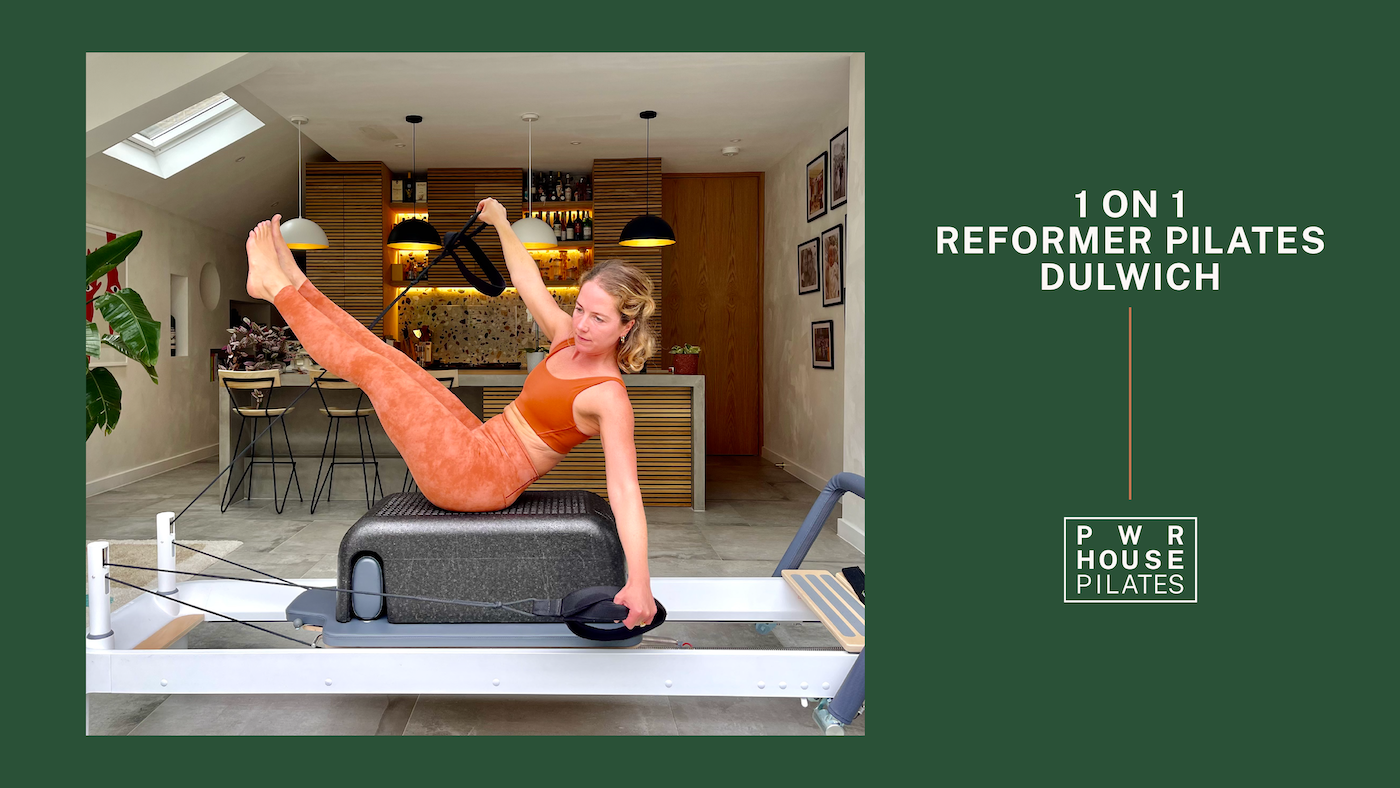 Woman performing reformer Pilates exercise on a Pilates reformer machine in a modern apartment with bar area and framed pictures on the wall.
