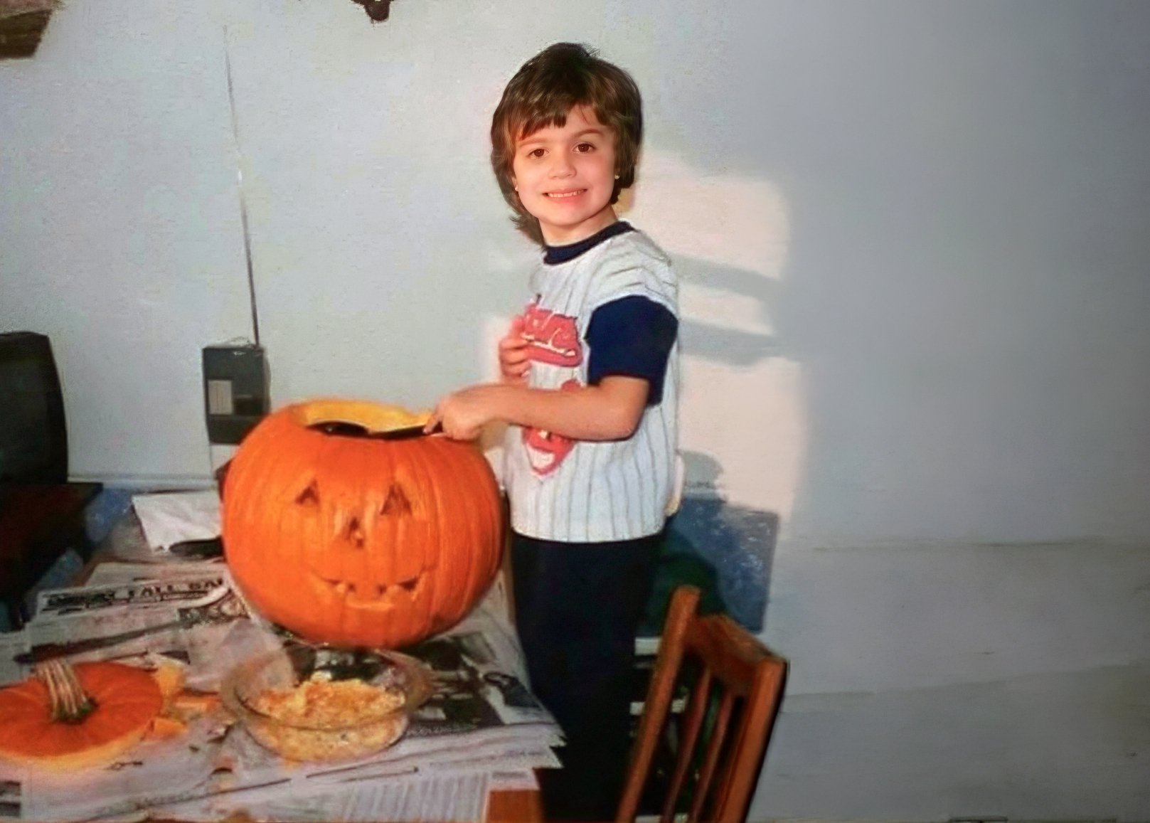 A young boy with brown hair smiling as he carves a face into a large orange pumpkin on a table.