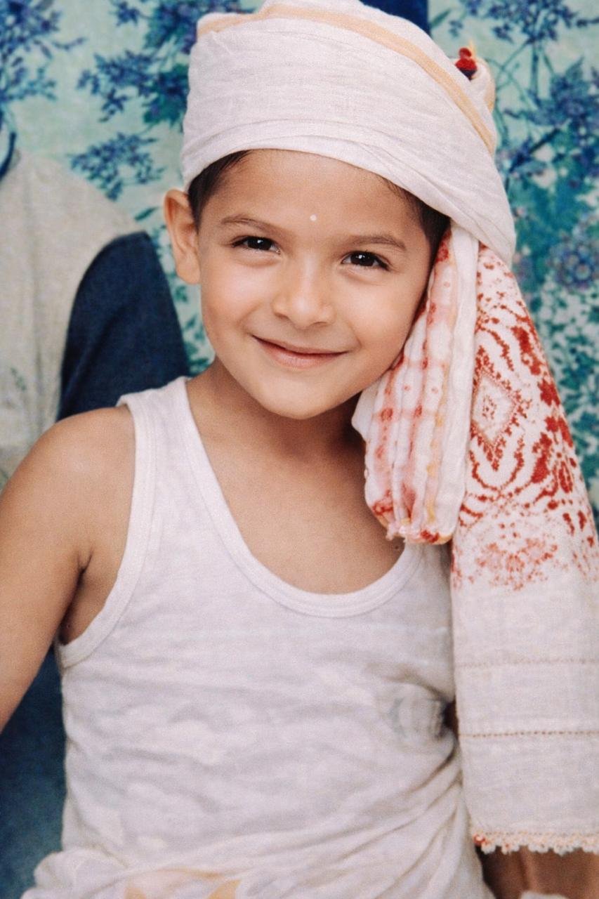 A smiling young boy with a white headwrap and a white sleeveless shirt, standing next to a woman in traditional attire, with a floral backdrop.