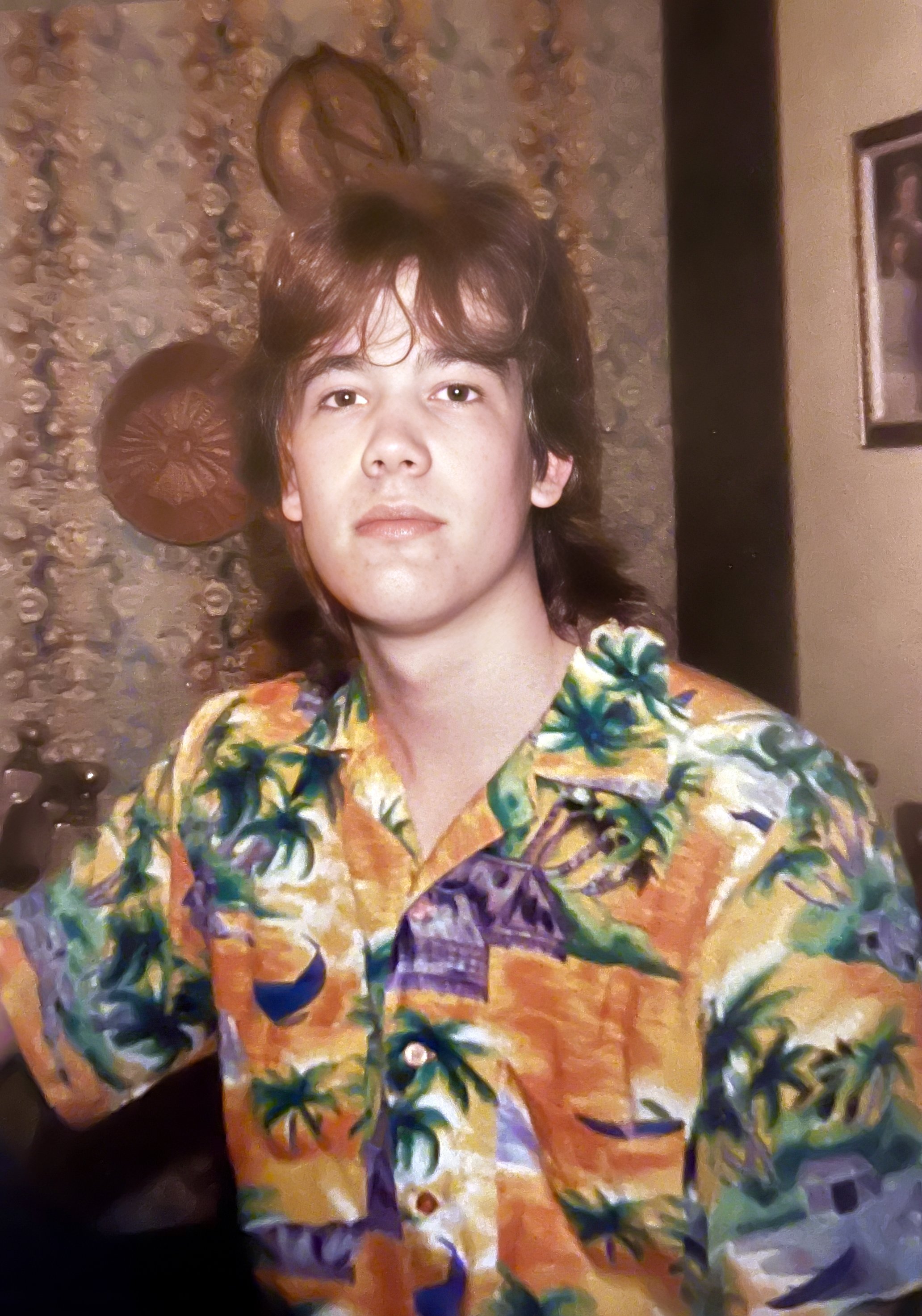 Young man with long, dark hair, wearing a colorful tropical shirt, sitting indoors in front of a textured wall with decorative plates and a framed photo.