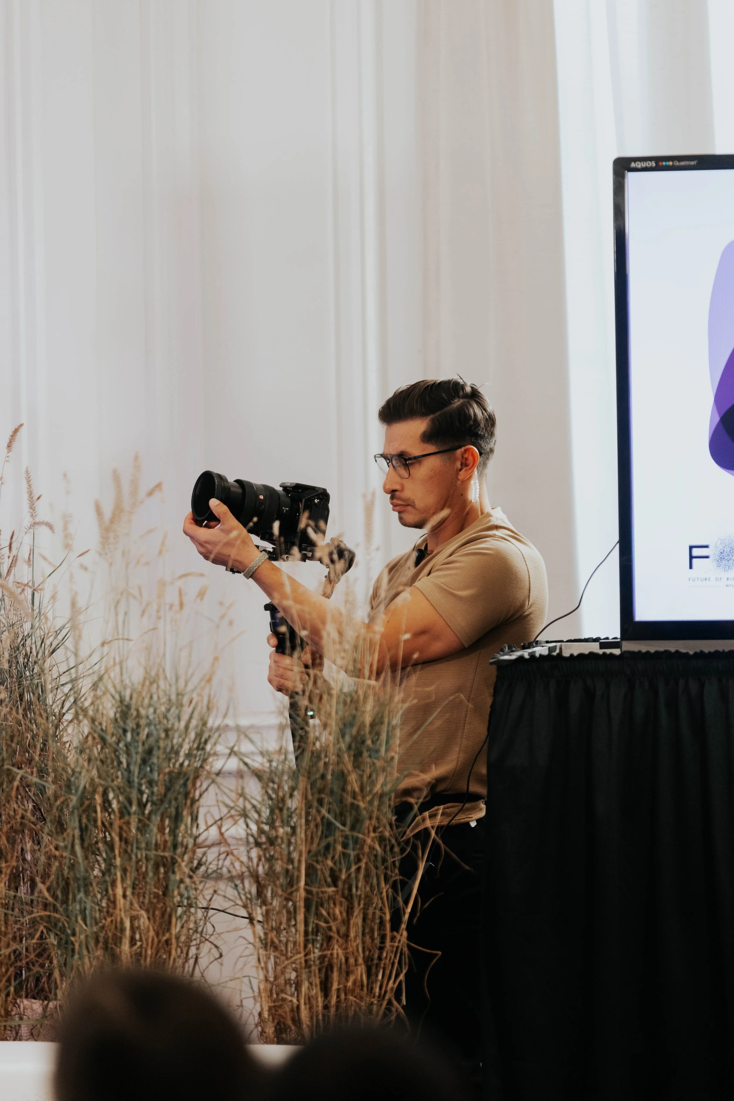 A man with dark hair and glasses setting up a camera on a tripod next to decorative plants at an indoor event.