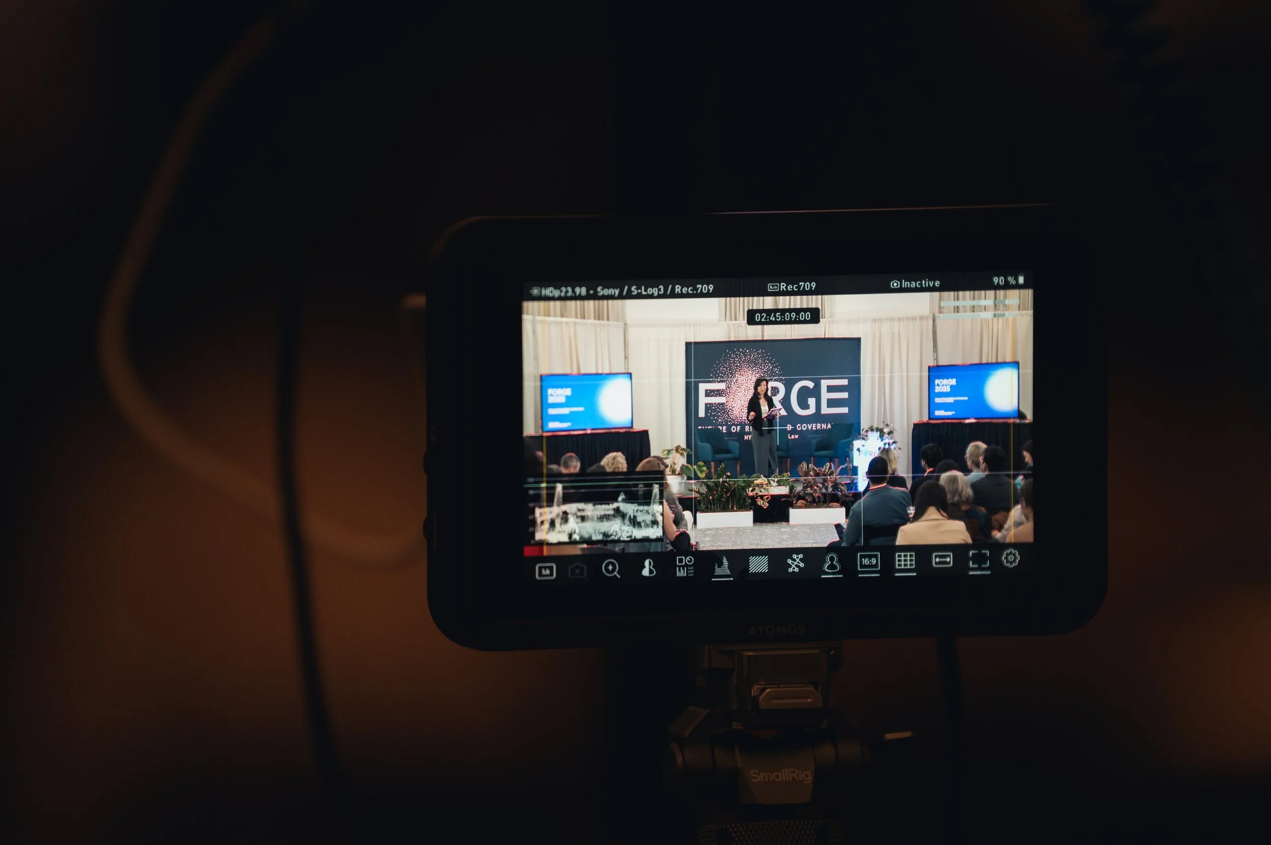 Video camera screen capturing a speaker on stage at a conference, with audience members seated and two presentation screens displaying "FORGE".