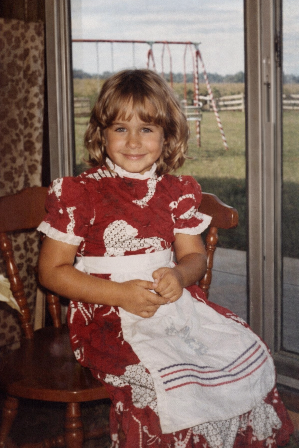A young girl with light brown hair and a bright smile is sitting on a wooden chair indoors. She is wearing a red dress with lace detailing and a white apron with colored stripes. Behind her is a sliding glass door revealing a backyard with a swing set and a wooden fence.