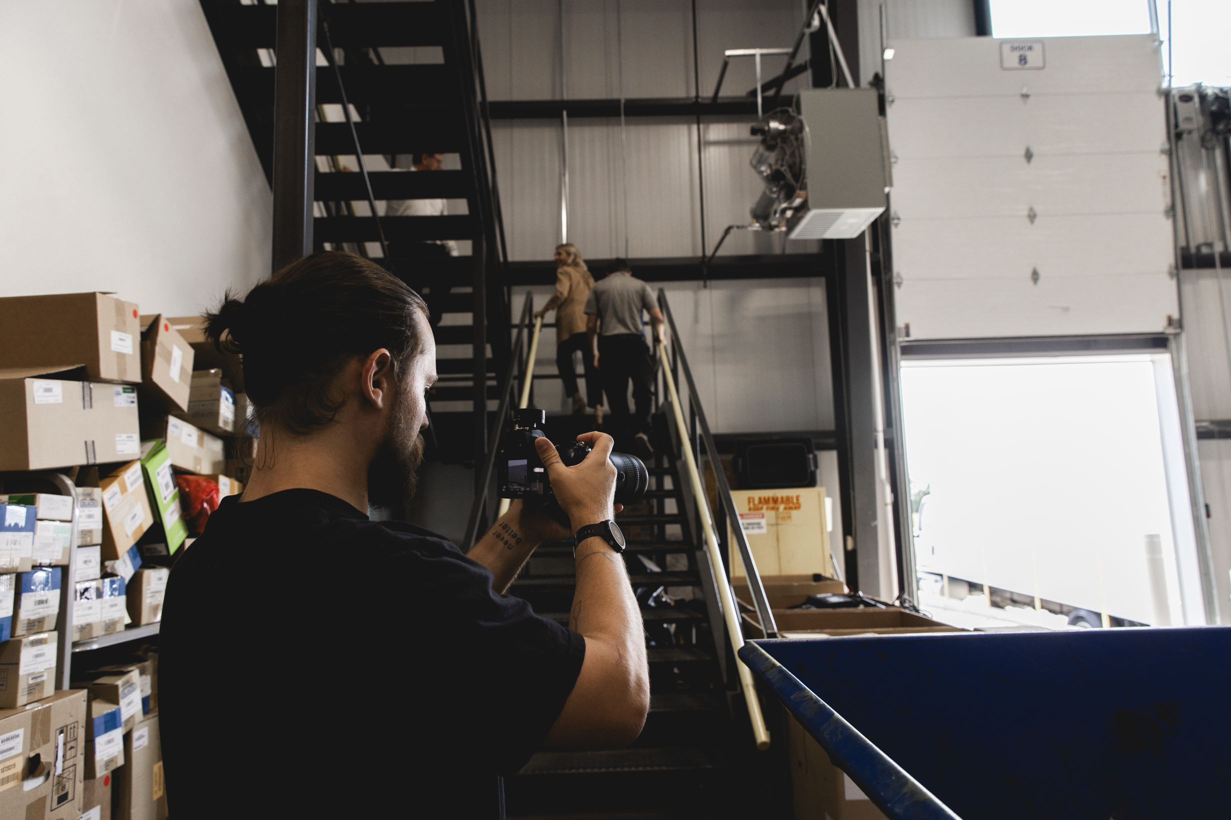 A man with long hair tied back, holding a camera, taking photos inside a warehouse. Two people are ascending a staircase in the background, near an industrial garage door.