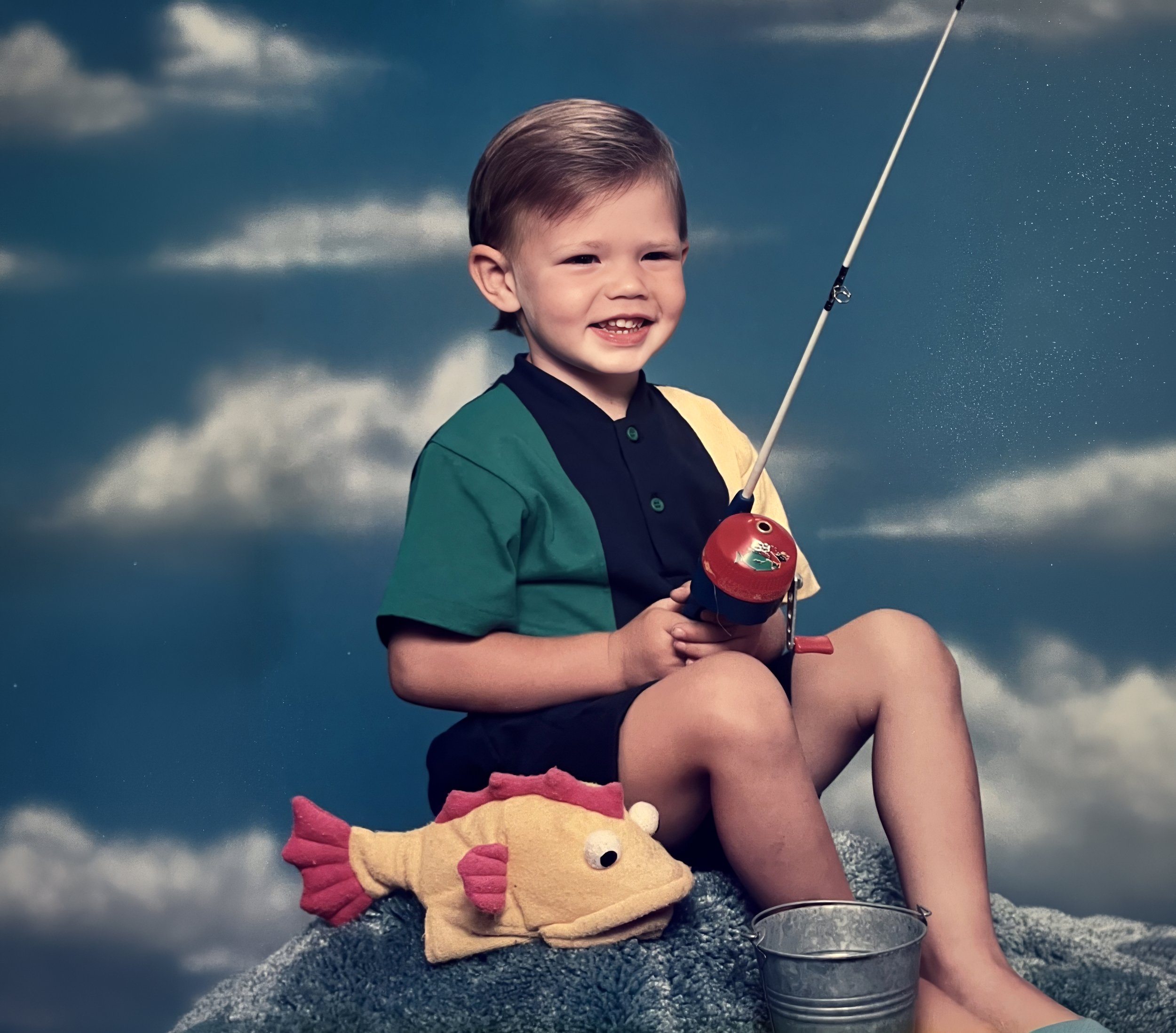 A young boy with short brown hair and a big smile sitting outdoors on a large rock, holding a fishing rod. He is wearing a colorful shirt and shorts, with a plush fish toy beside him and a metal bucket on the ground. The background features a cloudy sky.