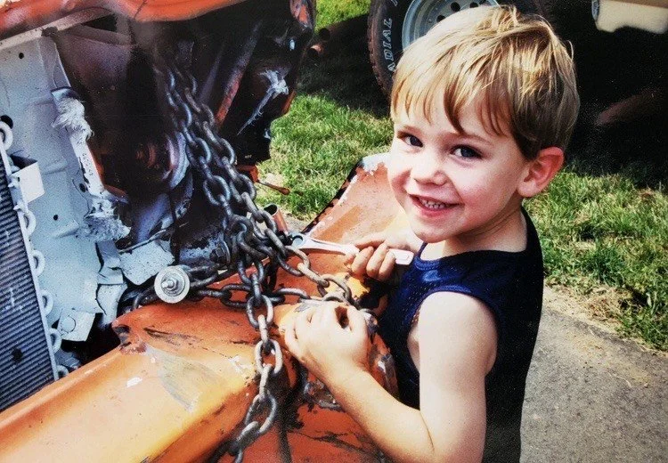 A young boy with blonde hair and a blue sleeveless shirt smiling while working on a tractor, holding chains attached to the tractor's front, outdoors with grass in the background.