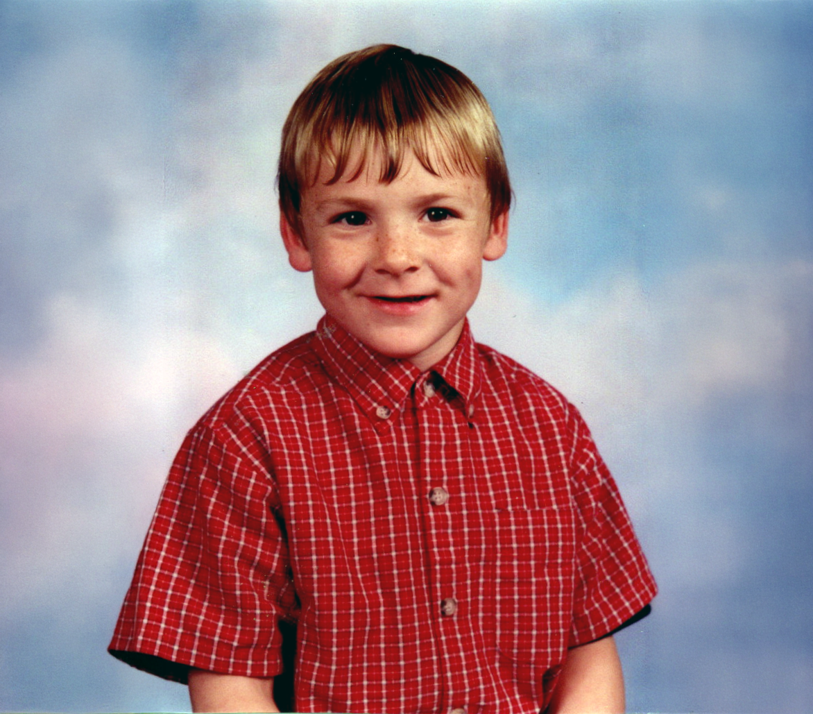 A young boy with blonde hair, freckles, and a red checkered shirt smiling at the camera, with a cloudy sky background.