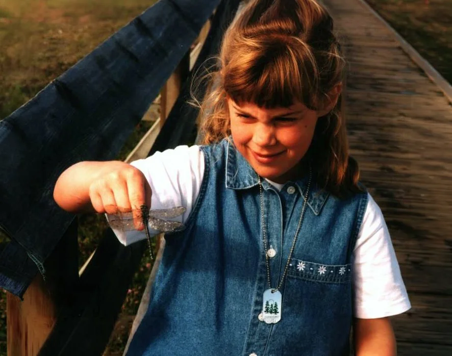 Young girl holding a dragonfly on her finger while standing on a wooden bridge outdoors