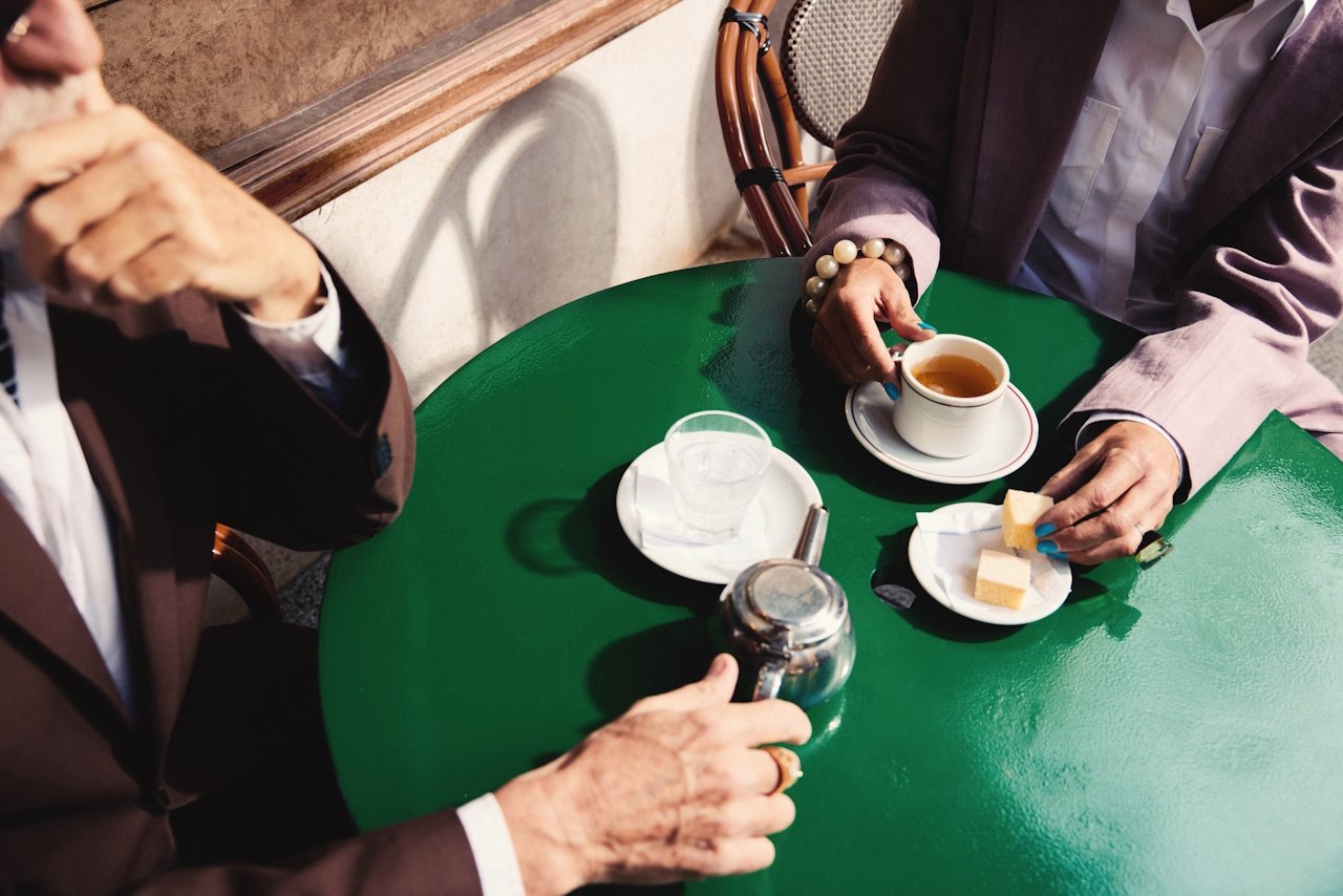 Two people sitting at a green table having coffee and snacks, with one person holding a sugar jar and the other holding a coffee cup and a plate with a piece of cake.