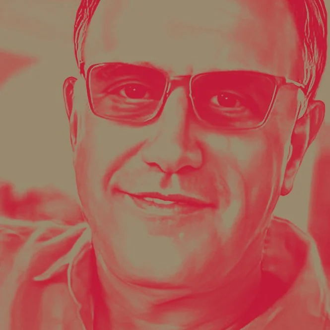 Close-up of a man wearing glasses, smiling at the camera with a blurred background.